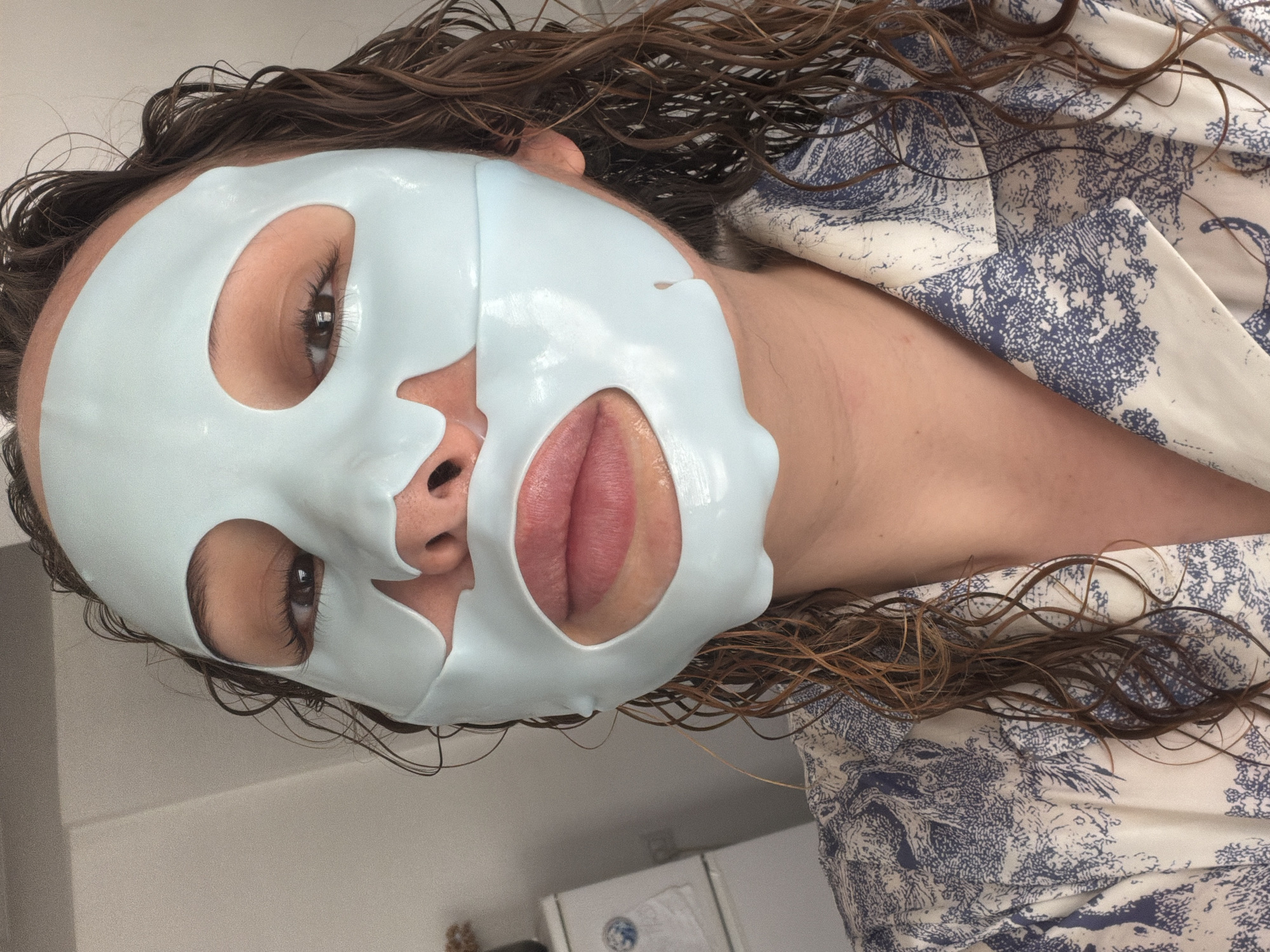 A person with wet, wavy brown hair is wearing a light blue sheet mask on their face. The mask has cutouts for the eyes, nose, and mouth. The person is wearing a white and blue patterned robe or top. The background is a plain, light-colored wall. The image was taken in Ramat Gan, Israel. The location within Ramat Gan is unspecified.