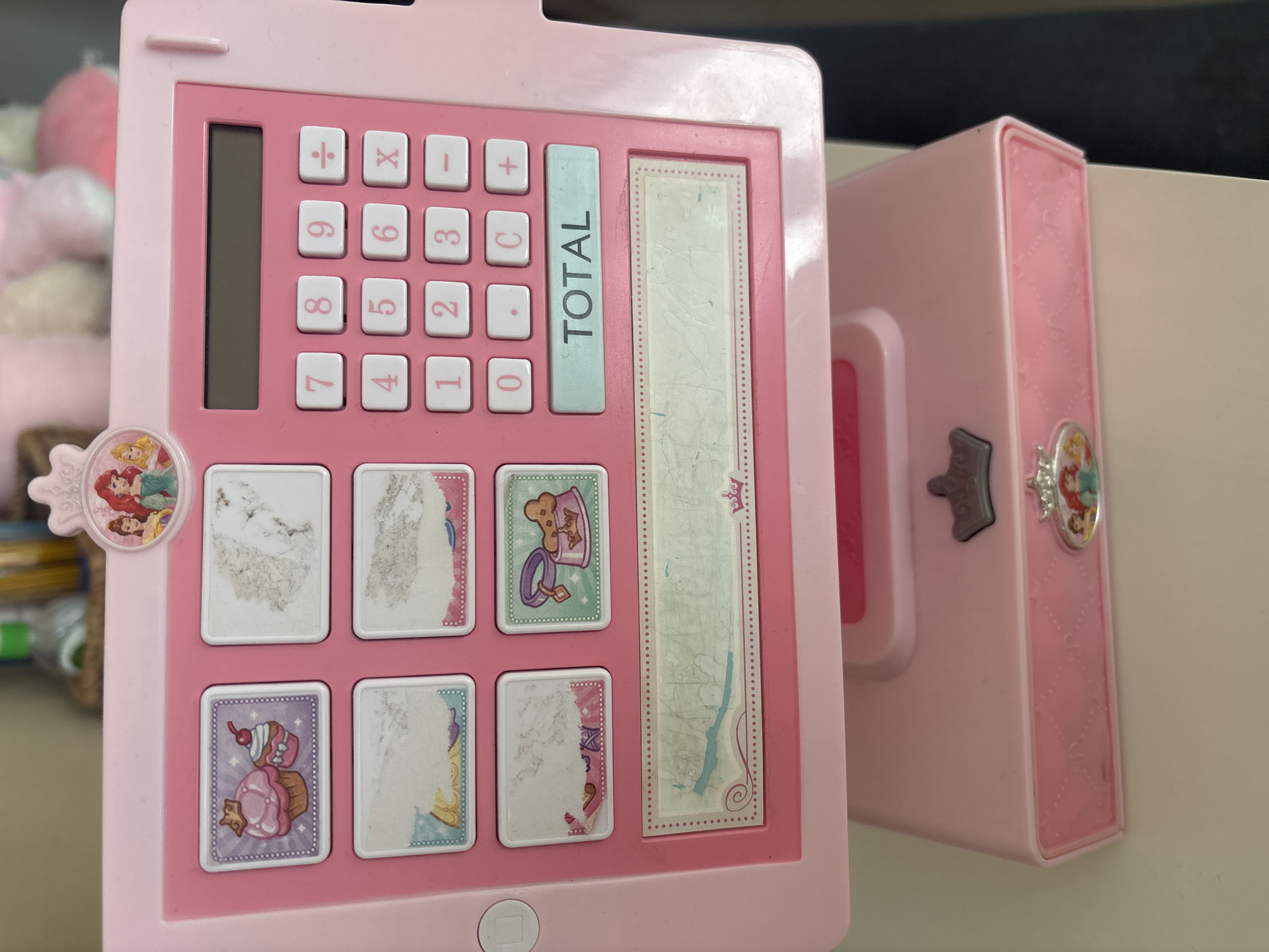 The image shows a pink toy cash register with a screen and a keypad. It is decorated with princess imagery, including Ariel and Cinderella. The cash register has several buttons with numbers and symbols, as well as a "TOTAL" button. There are also four square areas with pictures of cupcakes, a pet food bowl, and two blank areas. The cash register is open, revealing a drawer at the bottom. The scene appears to be indoors, with a blurred background that suggests a home or retail environment. No specific location in Dubai is discernible.
