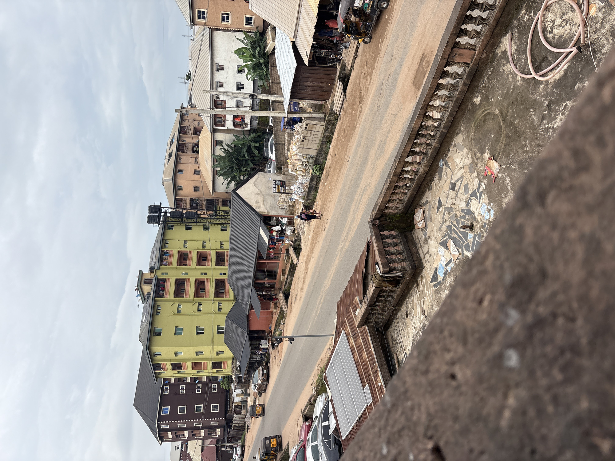 A street scene in Nkpor, Nigeria, is depicted under an overcast sky. The view is from an elevated perspective, looking down at a two-lane asphalt road. On the left side of the road, several cars are parked, including a silver sedan and a red sedan, along with three auto-rickshaws, two of which are yellow and one is dark colored. Further down the road, a white sedan is seen driving. Adjacent to the road, on the left, is a row of single-story shops with dark roofs. On the right side of the road, there are more shops and buildings. One building is a vibrant lime green, four stories high, with multiple balconies featuring orange railings. Another building behind it is dark brown, also four stories high, with white-framed windows. A group of people are visible walking on the right side of the road, and some are standing near the shops. Stacks of grey cement blocks are piled on the side of the road. A large pink hose is coiled on the ground in the foreground on the right. The foreground shows a weathered, ornate railing and a section of a roof with solar panels. The ground surface appears to be a mixture of dirt and broken tiles.