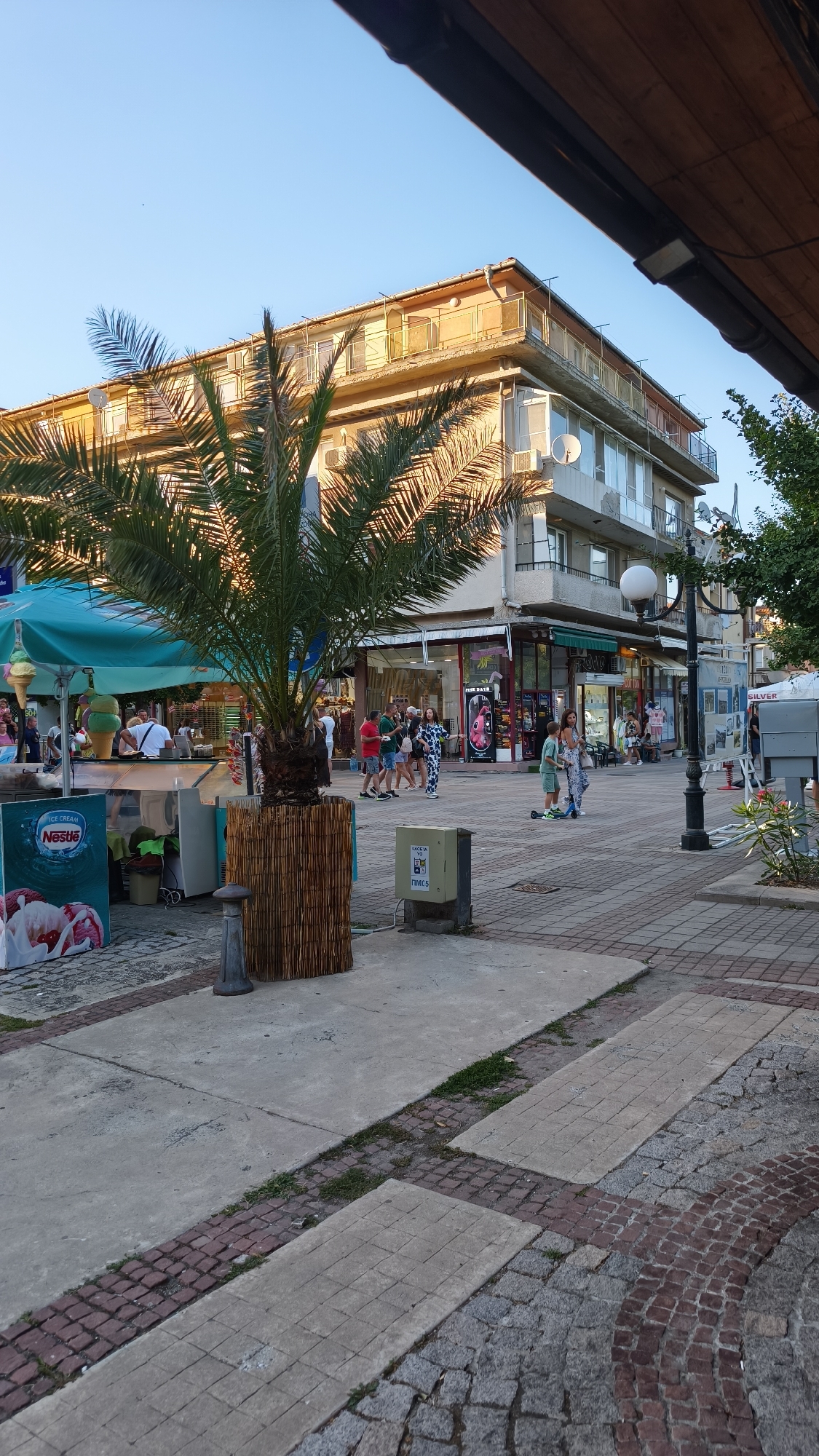 The image captures a street scene in Pomorie, Bulgaria, bathed in the warm light of a setting sun. The foreground features a concrete pathway interspersed with reddish-brown and grey cobblestones, leading towards a commercial building. To the left, a prominent palm tree in a woven planter stands out, with a Nestlé ice cream advertisement visible behind it. An ice cream stand with large cones is also present.
The commercial building is a multi-story structure with balconies and several shops on the ground floor. People are seen walking along the street, some browsing in the shops, while others are engaged in conversation. A few individuals are in motion, including a child on a scooter. The environment suggests a lively public space, likely a town square or a pedestrianized street. The sky is clear and blue.