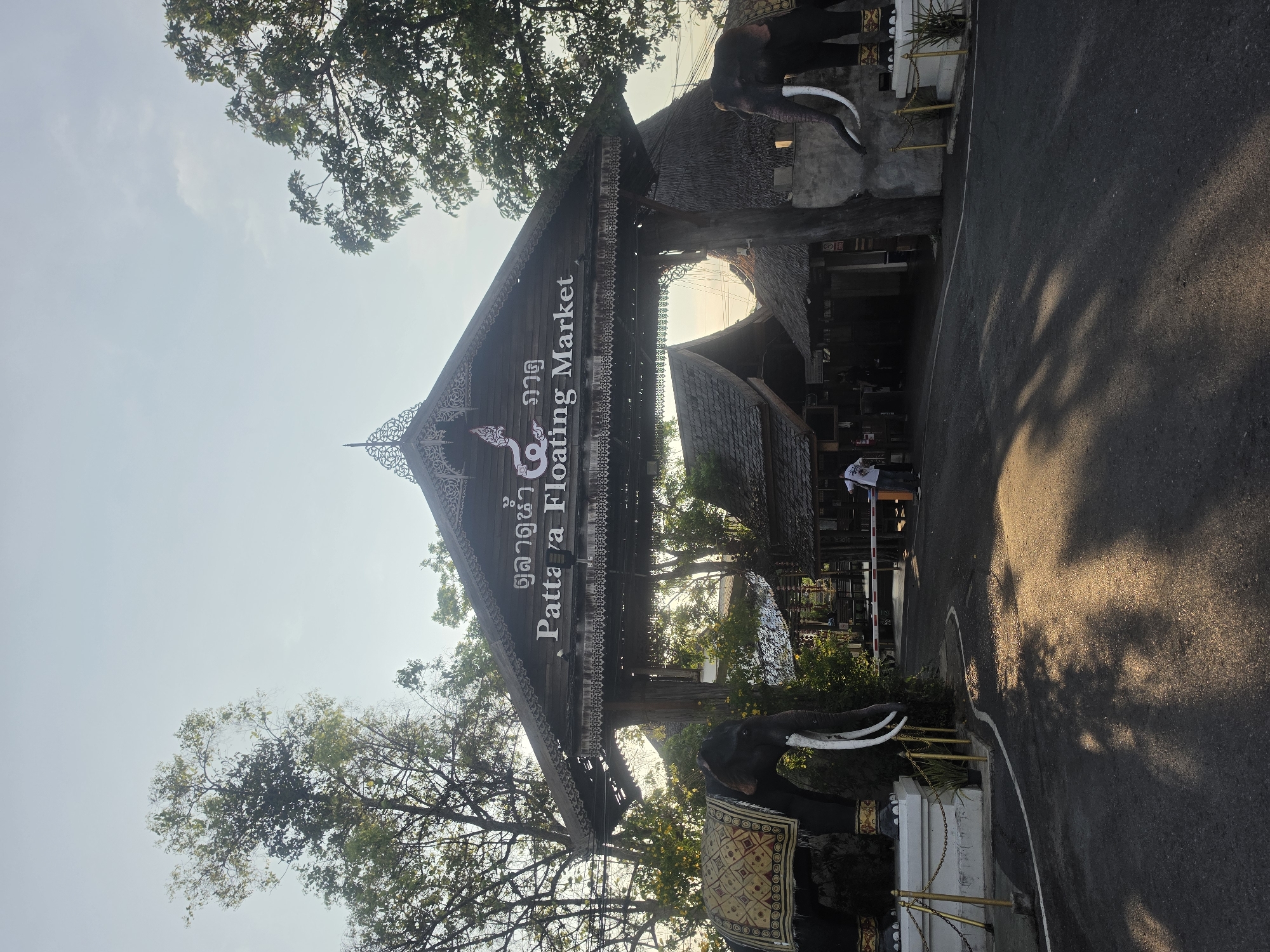 The image displays the grand, ornate entrance to the Pattaya Floating Market in Pattaya City, Thailand. A large wooden archway, featuring traditional Thai multi-tiered roofing and intricate carvings, dominates the center of the frame. White text on the dark wood fascia identifies the location, reading "ตลาดน้ำ ๔ ภาค" (Talad Nam Si Pak), which translates to "Four Regions Floating Market," alongside the English "Pattaya Floating Market."
Two large, dark elephant statues with long white tusks guard either side of the entrance. The elephant on the right is notably adorned with an elaborate gold and patterned fabric blanket. Lush green trees with dense foliage flank and arch over the entrance, contrasting with the bright, clear sky visible above.
An asphalt road or pathway leads into the market, with a distinct white line painted on its surface. Long, dark shadows, likely cast by the surrounding trees, stretch across the road, suggesting either early morning or late afternoon light. Beyond the main archway, additional traditional Thai-style buildings with thatched or tiled roofs can be seen, indicating the extent of the market complex. A few indistinct figures are visible further inside, suggesting visitors are present. The scene conveys a welcoming and culturally rich atmosphere typical of a popular tourist attraction.