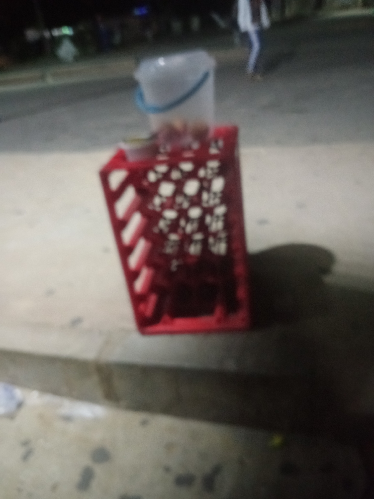 The image depicts a dimly lit outdoor scene, likely in Jalingo, Nigeria, focusing on a red plastic crate filled with white objects. A translucent white bucket with a blue rim sits atop the crate, also containing some of the white objects. The crate is positioned on a concrete curb next to a paved surface, possibly a street. In the background, the blurry outlines of buildings and lights suggest an urban or semi-urban environment at night. A person, also out of focus, is visible in the far background, walking away from the camera. The overall impression is of a street-side setup, possibly for a vendor or a temporary storage arrangement, with the contents of the crate and bucket appearing to be uniform and perhaps food-related.