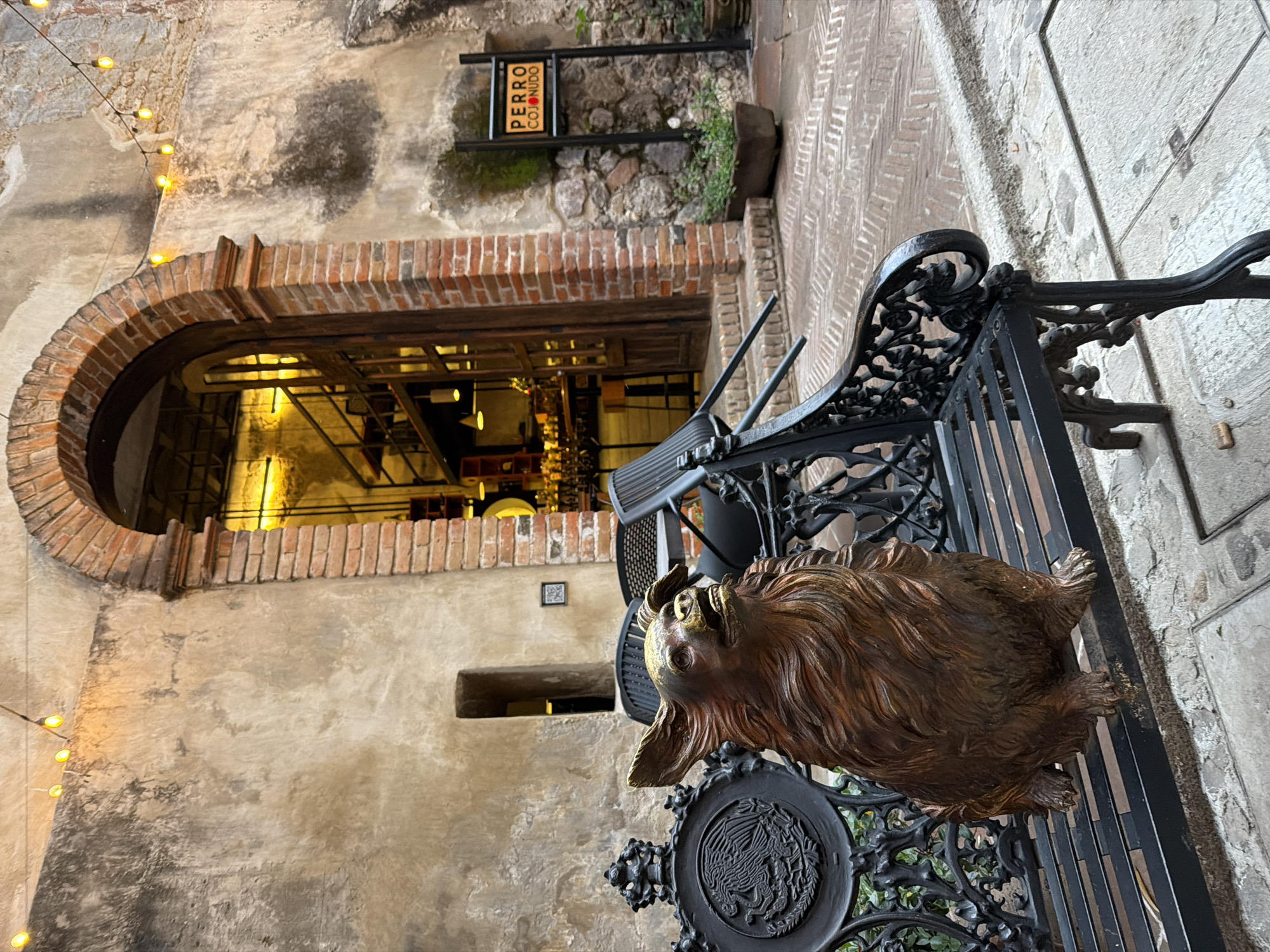 A medium-angle, rotated shot shows a street scene in what is identified as the city of Domingo Arenas, Mexico. The shot is focused on the architectural details of a building and surrounding elements.
On the left, an archway, constructed of red brick, leads into an interior space illuminated with artificial light. The architectural style is reminiscent of older construction, with rough plaster walls and exposed brickwork. A string of decorative lights hangs along the wall, adding a warm glow to the scene.
In the foreground, a decorative metal bench is positioned. A bronze-colored dog sculpture is attached to the bench. The sculpture is detailed, showing the dog's fur and features.
Behind the bench, a narrow street runs diagonally, showing cobblestone pavement. A sign with the words "PERRO COJONUDO" is attached to the wall, indicating a local establishment. The sign is surrounded by vegetation and is partially obscured by the angle of the shot. The wall surrounding the sign is made of stone.
The overall composition of the shot suggests a blend of old-world charm and modern elements. The scene is static, with no visible people or signs of movement. The setting appears to be a quiet street in the identified city.