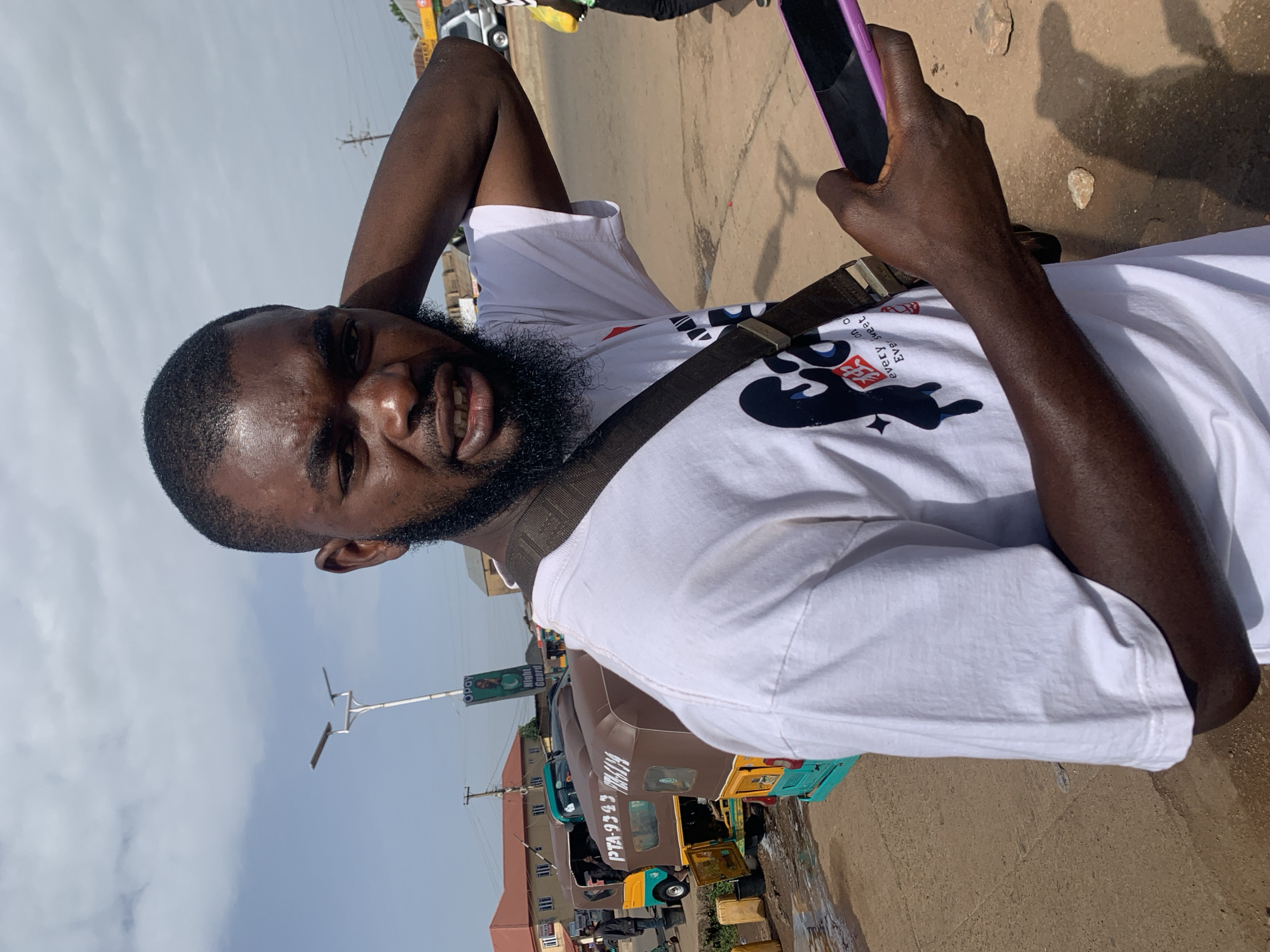 The image shows a close-up of a Black man, outdoors, with his arm behind his head. He is wearing a white t-shirt with a graphic design and a dark strap across his chest. He is holding a purple smartphone in his right hand. The man has a beard and is looking towards the camera with a slight smile.
The background suggests an outdoor setting, possibly a street or a market in Gura Top, Nigeria, given the context. There are vehicles, including a brown and green auto-rickshaw, and buildings visible in the distance. The ground appears to be made of dirt or gravel, with some scattered stones. The sky is visible and appears cloudy. The lighting suggests it is daytime, possibly late morning or afternoon. The overall mood seems casual and relaxed.
Visible text includes "PTA-9343" and what appears to be "KM 4134" on the side of the auto-rickshaw. There is also a green banner with the text "Night Guard" and "OPay" visible on a pole in the background. The graphic on the man's shirt includes stylized black figures.