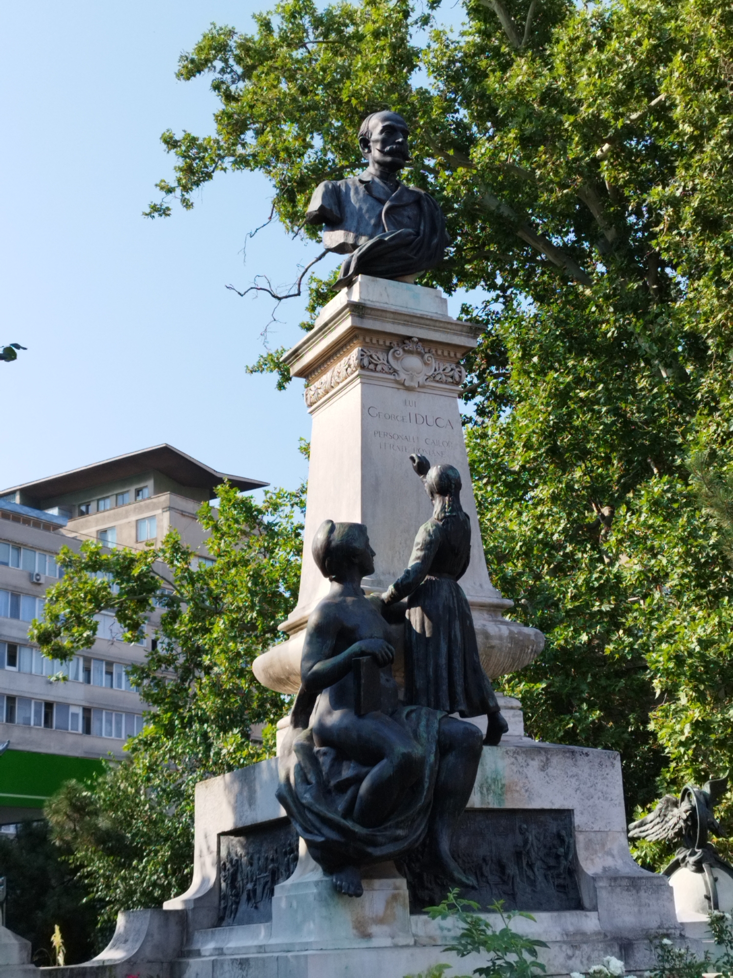 This image shows a statue in Bucharest, Romania, located in front of a modern building. The statue is a bust on a pedestal, with two smaller figures at the base. The bust depicts a man with a mustache, wearing a suit. The pedestal has inscriptions that are partially visible. The smaller figures are a seated woman holding a book and a standing woman pointing. The environment is sunny, with lush green trees and a clear blue sky. The modern building in the background appears to be an office or residential building with multiple windows.