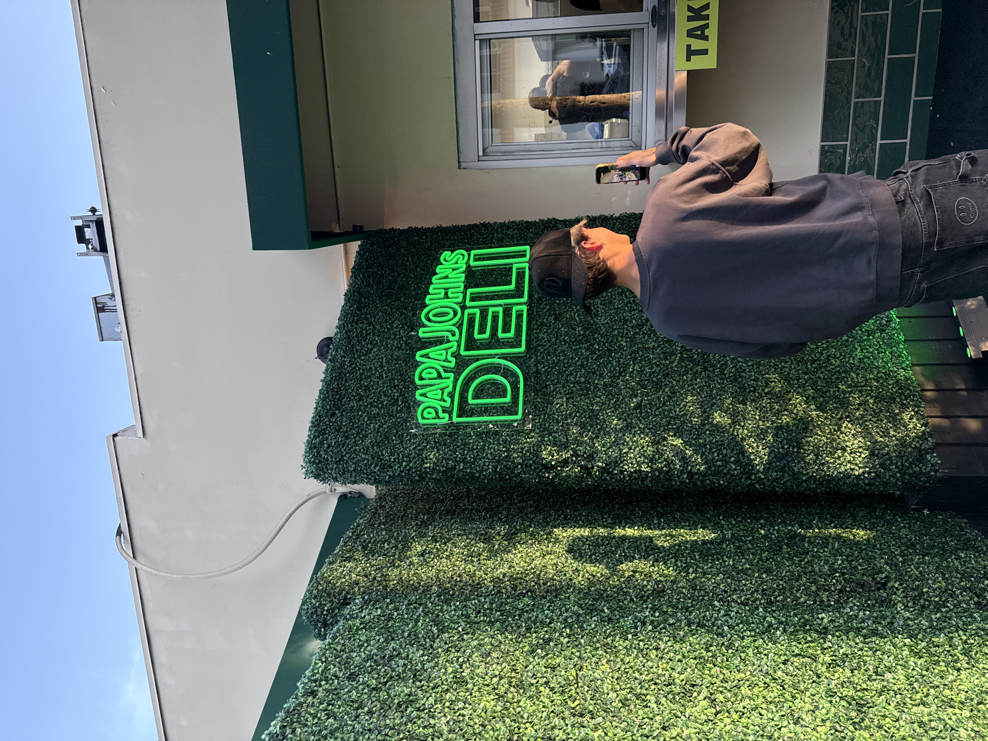 The image captures an outdoor scene at what appears to be a commercial establishment, likely in Los Angeles, USA, given the context. The photograph is oriented vertically, showing a building facade from a low angle under a clear blue sky.
The main subject is a man standing with his back partially to the camera, holding a smartphone in his right hand, seemingly taking a picture or video of the neon sign. He wears a dark long-sleeved sweatshirt, a dark baseball cap, and dark jeans with a distinct circular white logo featuring a smiley face and the word "drew."
The building features a large, dark green faux-hedge wall, creating a vibrant backdrop. Mounted on this hedge wall is a bright green neon sign that clearly reads "PAPA JOHNS DELI." Above the hedge, part of the building's light-colored wall is visible, along with a drive-thru or service window. Inside this window, a yellow sign with the partial word "TAKE" is visible, along with reflections showing part of the man and the environment.
The lighting suggests it is daytime, with clear sunlight casting shadows on the textured faux-hedge wall. In the upper left corner, a piece of equipment is mounted on the roofline. The overall aesthetic, with the neon sign and hedge wall, suggests a modern and possibly trendy establishment.