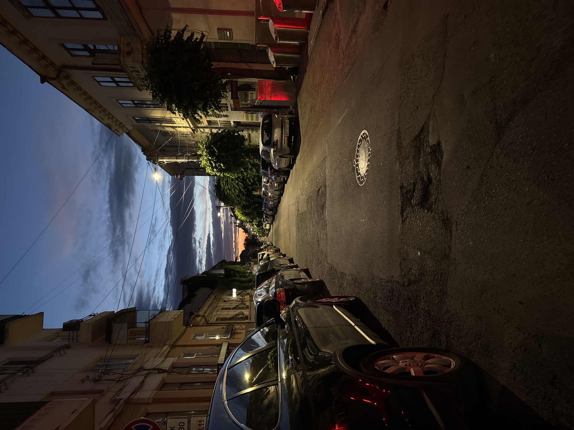 The image depicts a street scene in Chernivtsi, Ukraine, at dusk. The sky is a deep blue with dramatic clouds tinged with orange and pink hues from the setting sun. The street is lined with parked cars on both sides, creating a tunnel effect leading into the distance. Buildings with balconies and windows, characteristic of European architecture, flank the street.
In the foreground, a dark SUV is parked on the left side of the street, its reflective surfaces catching the dim light. The road surface is asphalt and appears somewhat worn, with a manhole cover visible in the middle of the lane. On the right side of the street, further down, are trash receptacles, one of which is illuminated with a red light. The overall atmosphere is calm and quiet, with no people visible in the scene. The lighting suggests the transition from daylight to evening.