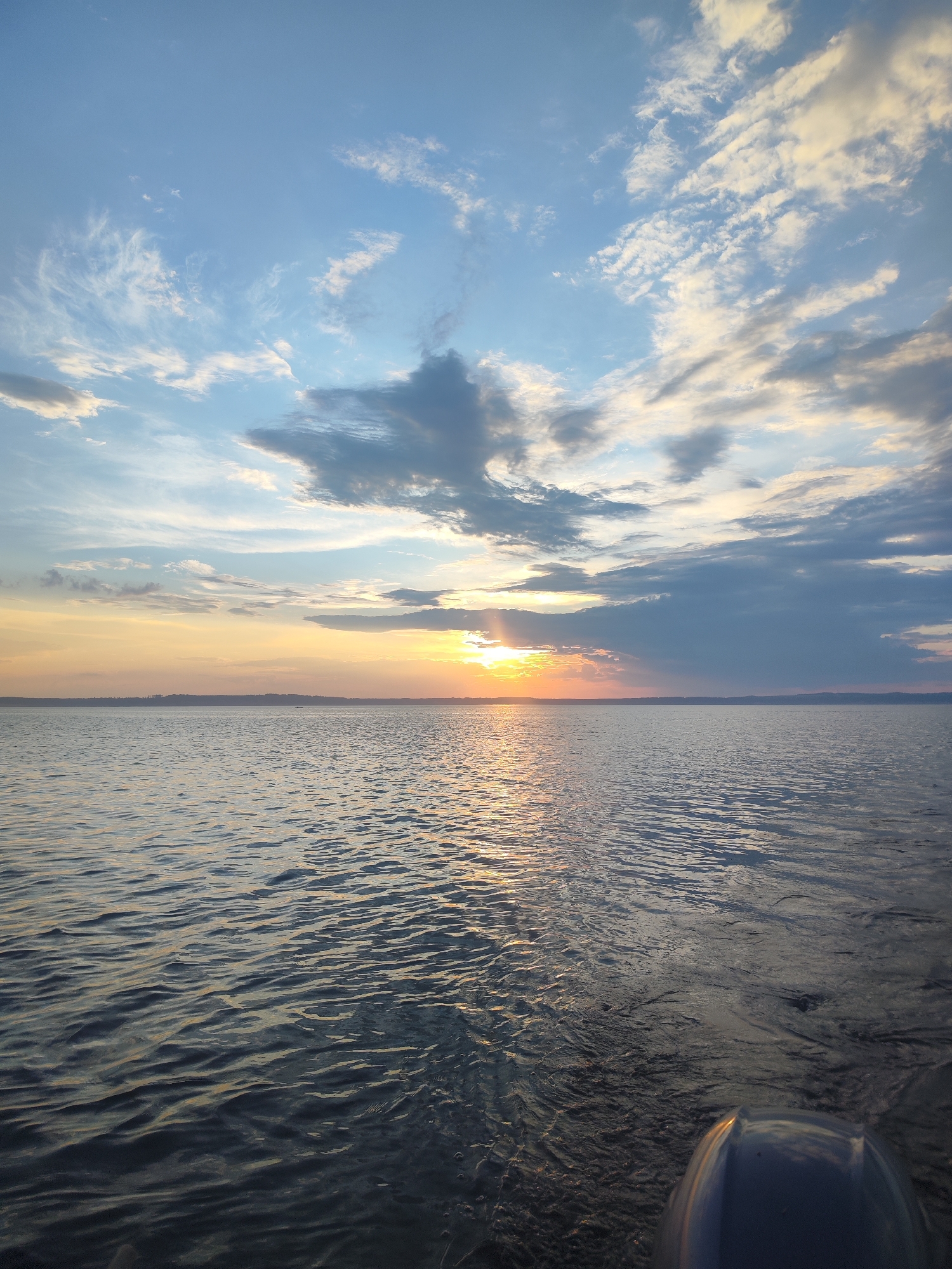 A wide-angle, eye-level shot captures a serene sunset over a large body of water, likely a lake or calm sea, in Galevo, Russia. The sky is a dynamic canvas of blues, oranges, and grays, with scattered clouds catching the warm glow of the setting sun. The sun itself is a bright, diffused orb peeking through a bank of clouds near the horizon, casting a shimmering golden path across the water's surface. The water is calm, with gentle ripples reflecting the sky's colors, and appears dark in the foreground, transitioning to a brighter, more reflective surface where the sun's rays hit. The distant shoreline is a faint, dark silhouette under the vibrant sky, suggesting a landmass or treeline. In the far distance on the water, a tiny dark speck may indicate a small boat or vessel. The lower right corner of the image shows the curved, dark surface of what appears to be part of a boat's hull or a similar structure, suggesting the photo was taken from a watercraft. The overall atmosphere is peaceful and expansive, emphasizing the vastness of nature.