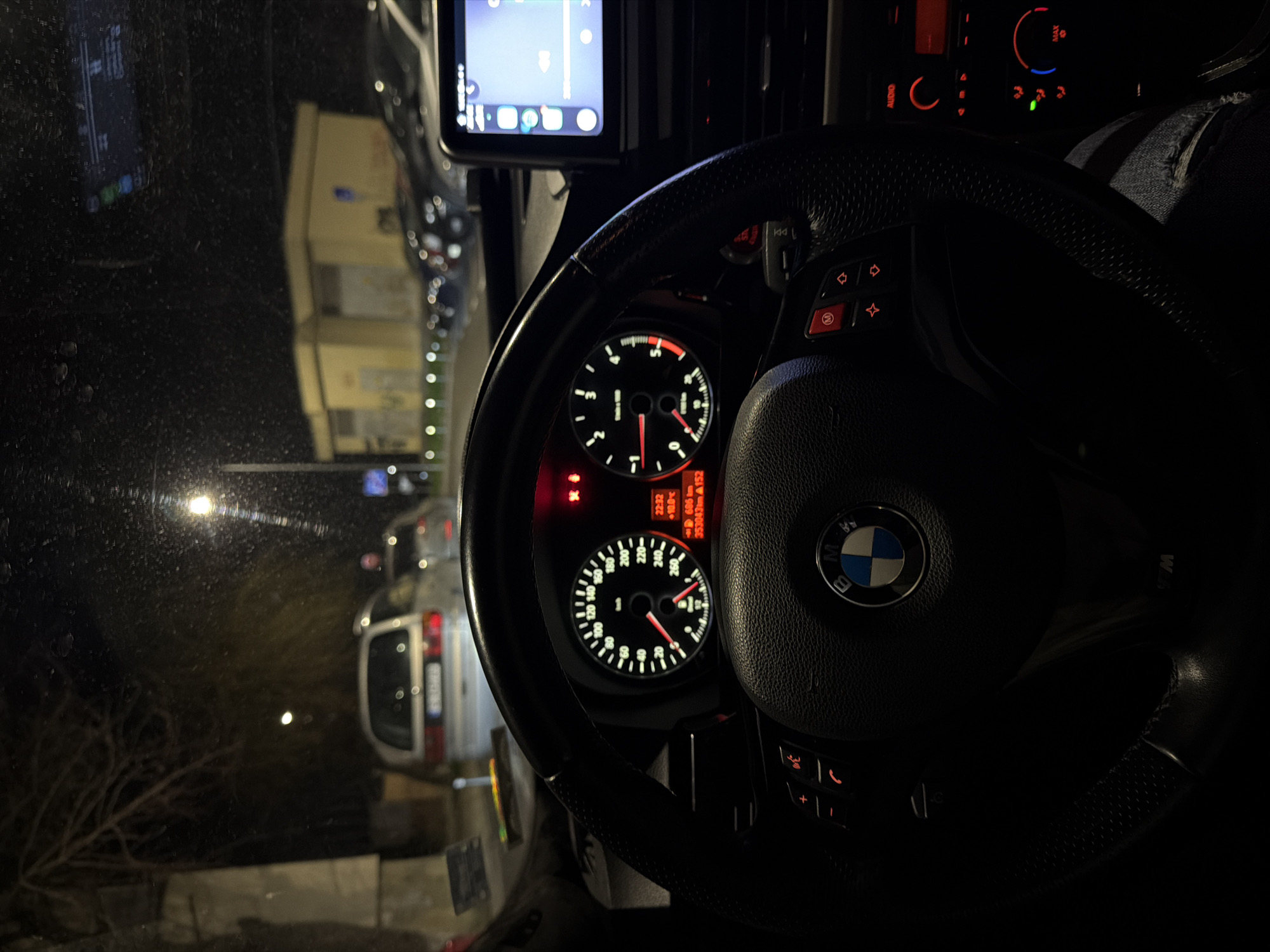 The image depicts the interior of a BMW vehicle, specifically from the driver's perspective, looking forward through a windshield partially covered in water droplets. It is nighttime. The steering wheel, black and leather-wrapped with an M-sport design, displays a central BMW logo and lower "M" badging, along with illuminated red controls for telephony and audio. The instrument cluster is illuminated, showing a speedometer on the left and a tachometer on the right, both with red needles. A digital display indicates the time as 22:32, an outside temperature of +10.0°C, a trip distance of 686 km, and a total odometer reading of 353043 km. The hazard warning lights indicator is illuminated. To the right, a mounted tablet-style infotainment screen displays a menu of applications, likely a smartphone mirroring interface. Below it, red-backlit climate and audio controls are partially visible. The driver's leg, clad in blue ripped jeans, is visible in the lower right.
Outside the vehicle, the scene is a street in Varna, Bulgaria, indicated by a partially visible Bulgarian license plate starting with "B" (Varna province code) on a silver minivan parked directly ahead. Several other dark-colored vehicles are parked on the right side of the street. A prominent light yellow, single-story building with a flat roof stands behind the parked cars on the right. Streetlights illuminate the environment, and bare tree branches are visible on the left side of the frame.