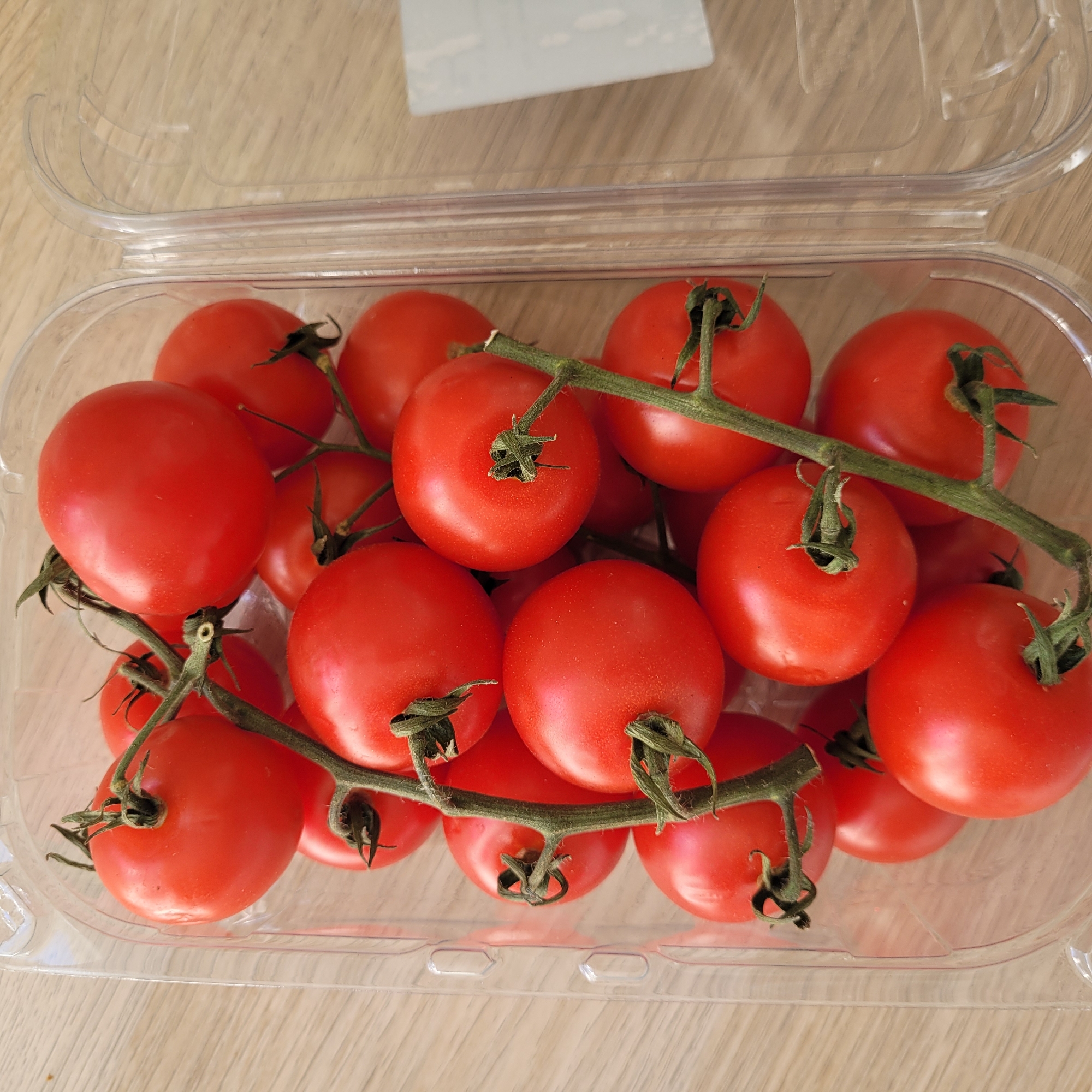The image shows a clear plastic container filled with ripe, red cherry tomatoes on the vine. The tomatoes are clustered together, with their green stems and calyxes visible. The container is sitting on a light-colored wooden surface. There are no people visible in the image. The scene is indoor and appears to be a domestic setting.