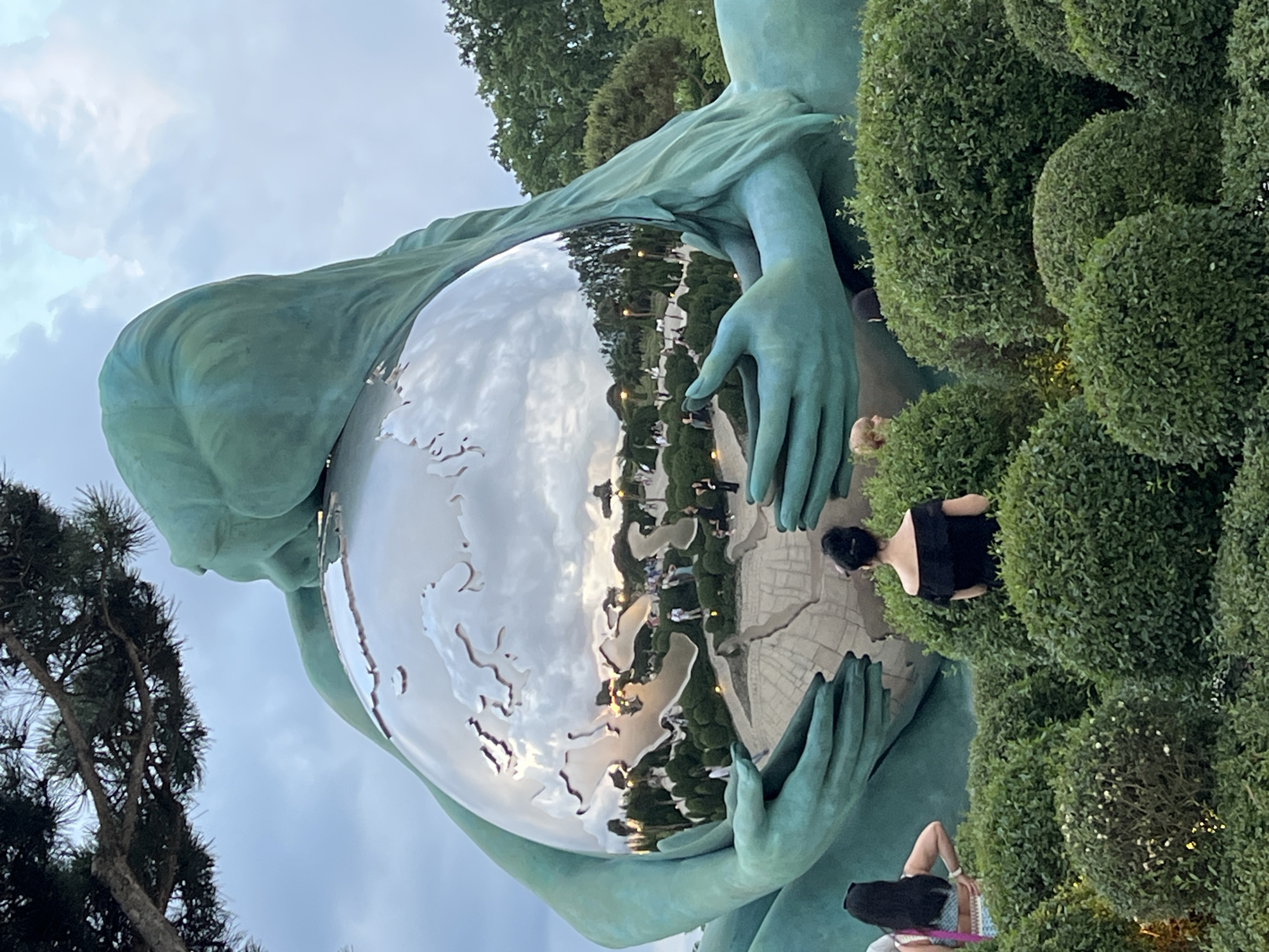 The image depicts a large sculpture of a woman holding a globe in her arms. The sculpture is made of a green material and the globe is made of a reflective, metallic material. The globe reflects the sky, clouds, and surrounding landscape, which includes manicured bushes and trees. Several people are visible in the background, some standing and some walking. The overall scene appears to be an outdoor public space, likely a park or garden. This sculpture is located in Krasnodar, Russia, specifically in the "Galitsky Park" (also known as the "Krasnodar Stadium Park"). The sculpture is titled "The Woman Holding the Earth."