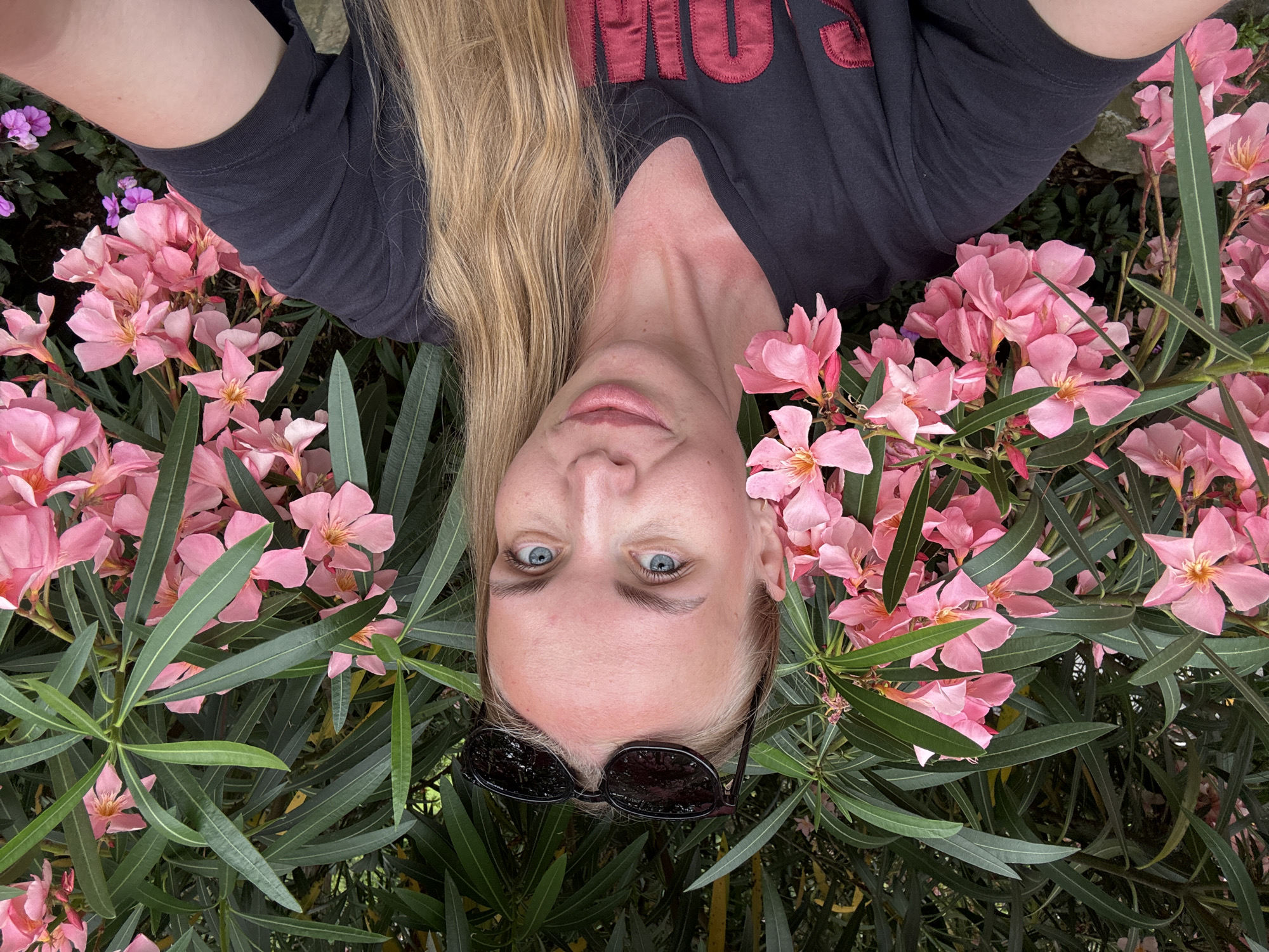 The image shows a young woman lying down, surrounded by pink oleander flowers and green foliage. Her head is positioned at the bottom of the frame, and her long blonde hair cascades around her. She is wearing a dark t-shirt with red lettering on the back. Sunglasses are perched on her forehead. Her blue eyes are open, and she has a neutral expression. The scene appears to be outdoors, with lush vegetation suggesting a garden or natural setting. The lighting is soft, indicating it could be daytime. The presence of oleander flowers suggests a warm climate. The image is oriented such that the woman is upside down relative to a typical upright perspective.