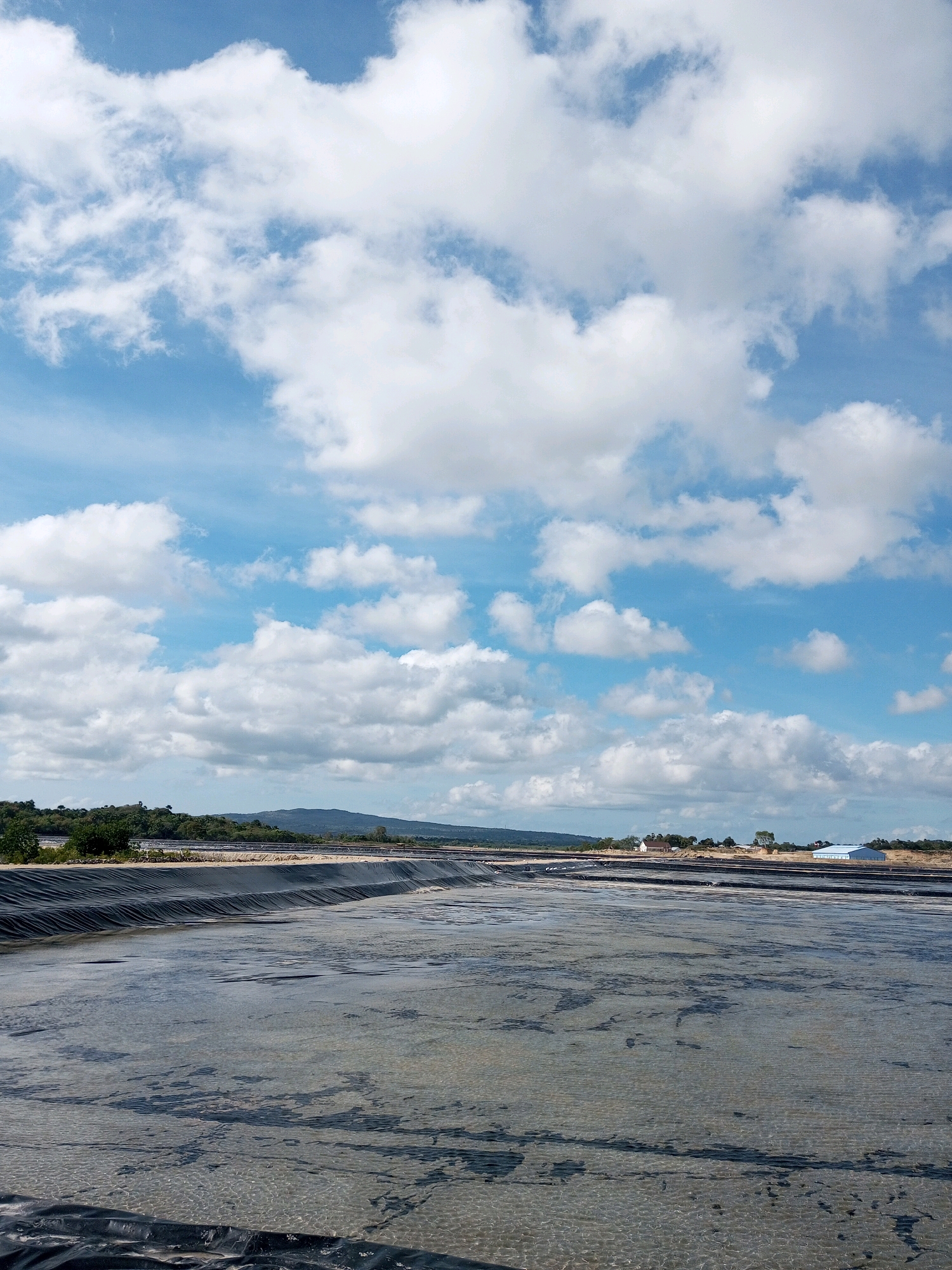 This is a vertical, eye-level, full shot of an outdoor industrial area on a bright, overcast day. In the foreground, there is a large, shallow body of water that appears to be a salt evaporation pond or similar industrial holding tank. The bottom of the pond is visible through the clear, shallow water, showing a textured, sandy surface with darker patches of what might be salt deposits or algae. The edges of the pond are lined with black plastic sheeting.
In the mid-ground, there are more of these lined ponds, stretching horizontally across the frame. Beyond the ponds, a line of green trees and vegetation runs along a low, rolling hillside. In the distance, the sky is a vivid blue, filled with numerous white, puffy cumulus clouds. On the right side of the mid-ground, a large, white, modern industrial building with a blue roof is visible. There are no people in the image. The location is specified as Serubeba, Indonesia.