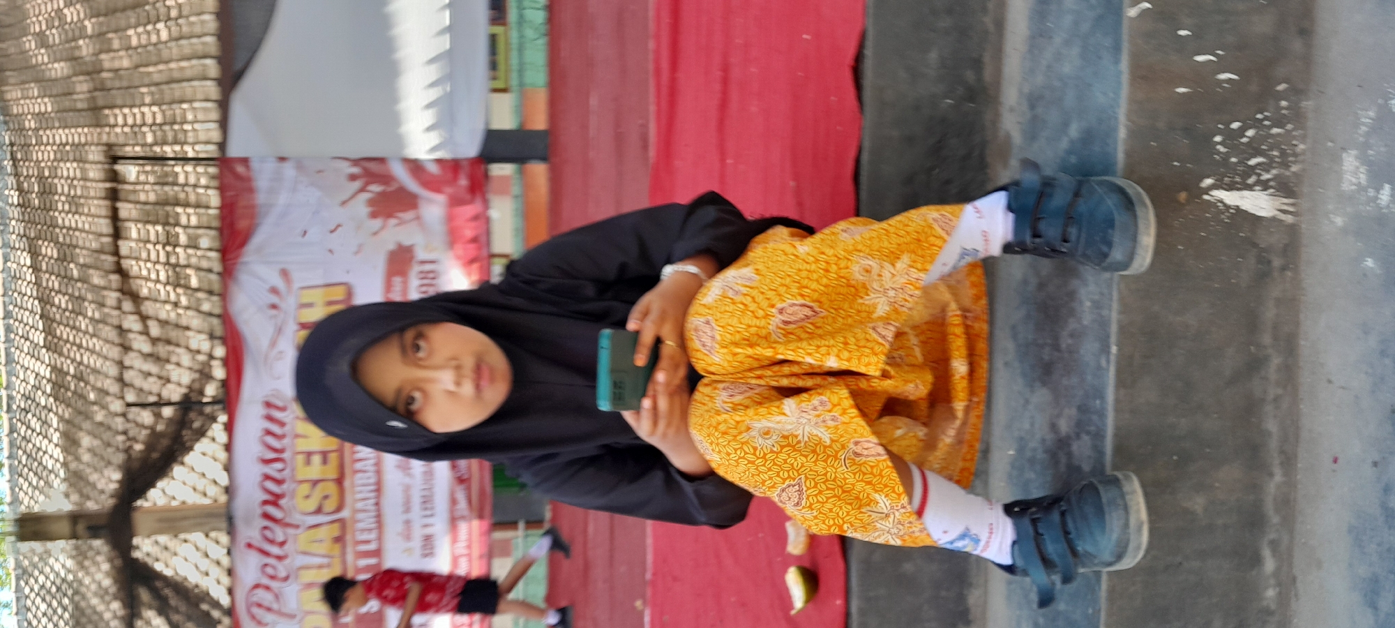 A young girl in a black hijab and a yellow patterned skirt sits on concrete steps in front of a red carpet. She holds a dark green smartphone in her hands, looking at it. Her white socks have red and blue stripes. A portion of a banner with Indonesian text, including "PELEPASAN" and "SEKOLAH," is visible behind her, suggesting the location is a school in Lemahbang Dewo, Indonesia. A slice of lime is on the red carpet to her left. Another child in a red shirt and dark shorts is blurred in the background. The roof of a building is visible above the banner.