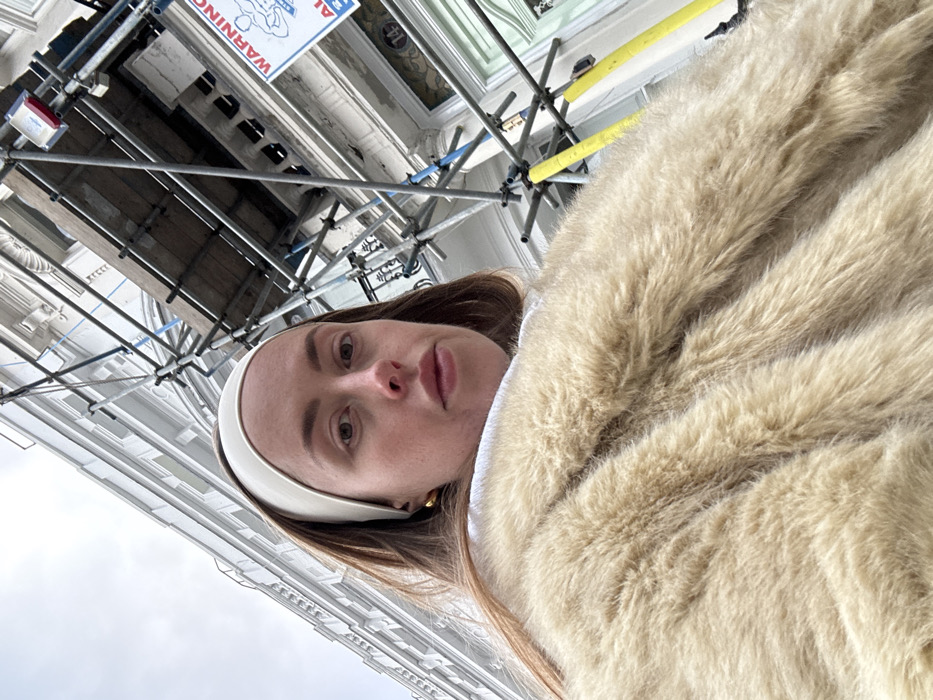 A low-angle, close-up photograph captures a female individual on a street in London, United Kingdom, beneath an overcast sky. The subject, occupying the central and lower portions of the frame, has light brown hair, fair skin, and wears a cream-colored headband and gold-toned hoop earrings. She is dressed in a substantial, light beige faux fur coat over a white garment, and her gaze is directed neutrally towards the camera. Behind her to the left, a multi-story, white stuccoed terraced building with classical architectural elements rises into the grey sky. To her right, an adjacent building section is covered by extensive metal scaffolding, featuring wooden planks and yellow protective sheaths on vertical poles. A partially visible white warning sign, displaying a black silhouette of a muscular figure, is attached to the scaffolding. Below the scaffolding, a light green entrance door with decorative glass panels is visible, with the house number "14" partially discernible above it.