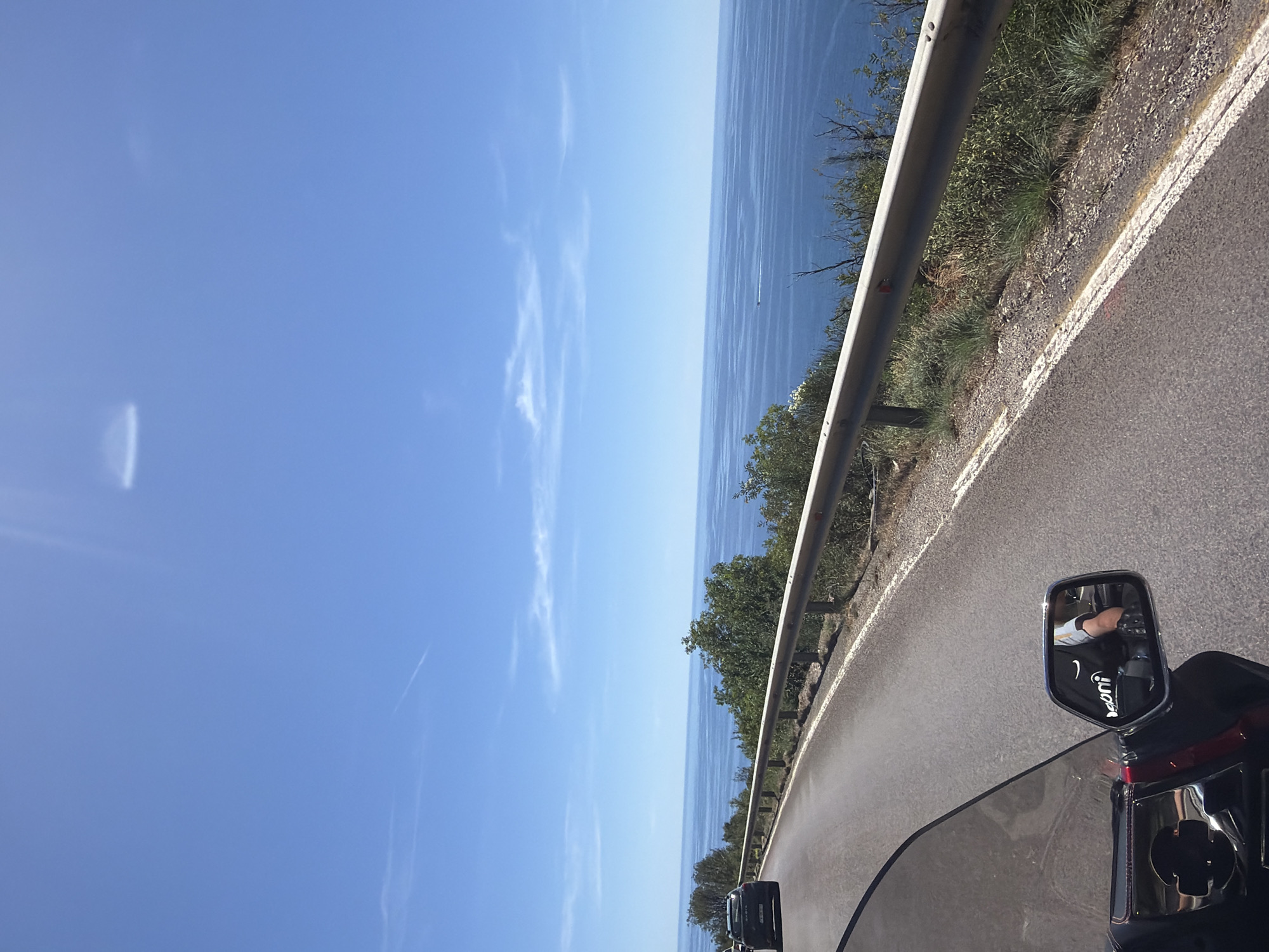 The image shows a view from a motorcycle traveling along a coastal road. The road is paved and curves to the left, with a metal guardrail along the right side. Beyond the guardrail, there are green trees and shrubs. The road overlooks a vast expanse of blue ocean, with a few small whitecaps visible. The sky is a clear, bright blue with some wispy white clouds. In the foreground, a portion of the motorcycle's windshield and a side mirror are visible. The side mirror reflects the image of a person's arm wearing a black glove and a white and yellow shirt. A dark colored car is seen on the road ahead. The scene appears to be in a sunny, coastal location.
