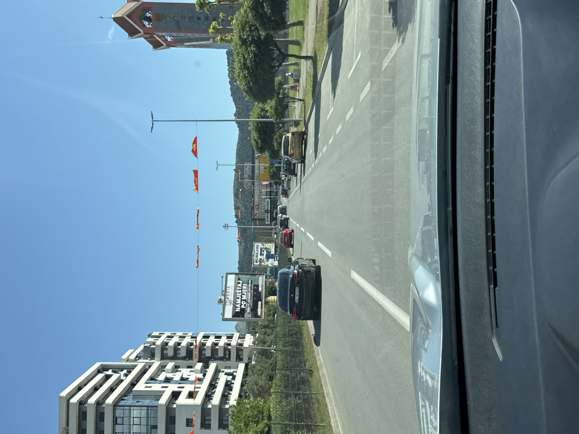 View from inside a car, looking forward down a street in Bar, Montenegro. The street has two lanes going in each direction, with dashed white lines separating the lanes. There are cars ahead in both lanes. On the left side of the road, there is a modern white building and a billboard. Further ahead, there are red banners hanging over the street. A tower is seen in the distance. The sky is clear and blue. The car's interior is visible on the right.