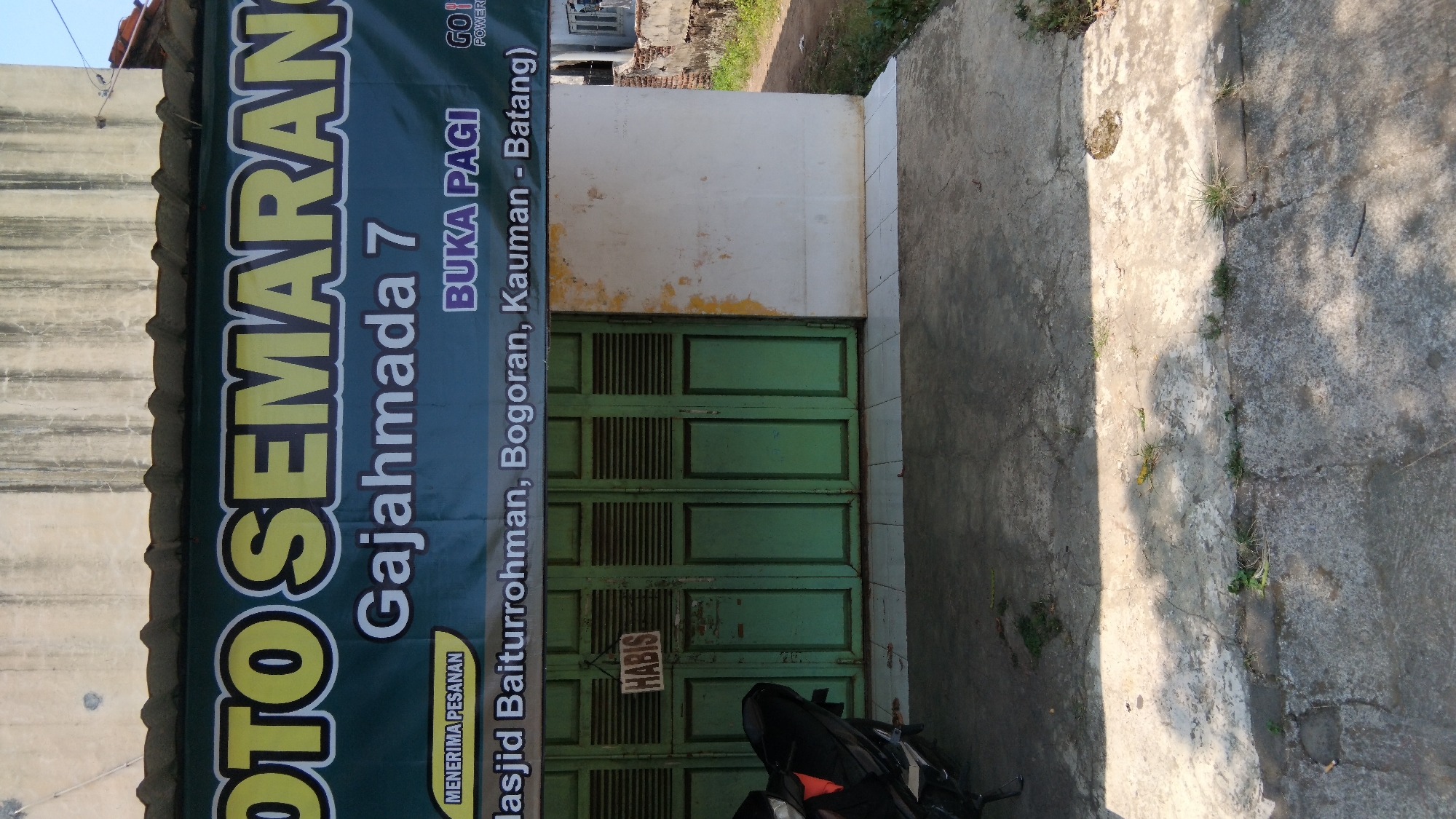 The image shows the exterior of a shop with a green facade and a banner above it. The banner displays "OTO SEMARANG" in large yellow letters with a black outline. Below that, it reads "Gajahmada 7" and "BUKA PAGI". To the left of "BUKA PAGI", a yellow banner states "MENERIMA PESANAN". The bottom of the banner has text in white, which appears to list locations: "Masjid Baiturrohman, Bogoran, Kauman - Batang)". A small logo with "GO!" and "POWER" is visible on the right side of the banner.
The shop entrance is covered by two large, weathered green doors, each with horizontal slats. A small sign that says "HABIS" (meaning "finished" or "sold out" in Indonesian) is hanging on the left door. To the left of the entrance, a portion of a black motorcycle is visible. The building facade above the banner is a muted color with visible weathering and stains. The ground in front of the shop is a paved area with cracks and some sparse vegetation growing through. Sunlight casts shadows on the ground. The overall scene suggests a small, local business in Indonesia. While the banner mentions "Batang", which is a regency in Central Java, the prompt specifies the location as "Proyonangg, Indonesia". Without further information or context, it is not possible to definitively confirm the location as Proyonangg based solely on the visual information provided.