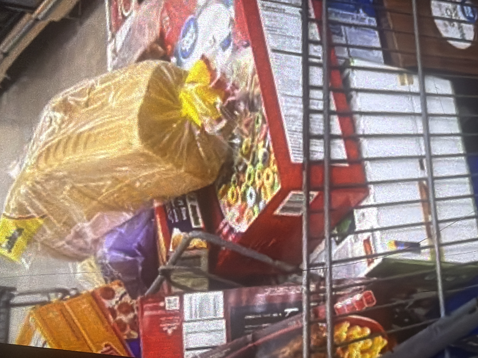 The image depicts a close-up, somewhat blurry view of groceries inside a shopping cart, likely at a store. In the foreground, a loaf of bread wrapped in clear plastic is prominent, with a yellow twist tie securing the bag. Behind the bread, several items are stacked: a box with a colorful, patterned design (possibly cereal or snacks), a red box with a white label, and a purple plastic bag. Further back and to the left, a red box with some text and graphics is visible, and above that, a red dish containing yellow food items. The background shows the metal bars of the shopping cart and other indistinct items on shelves. The overall lighting is bright, with some reflections on the plastic packaging. The image does not provide any direct visual clues to its specific location in Bukuru, Nigeria.