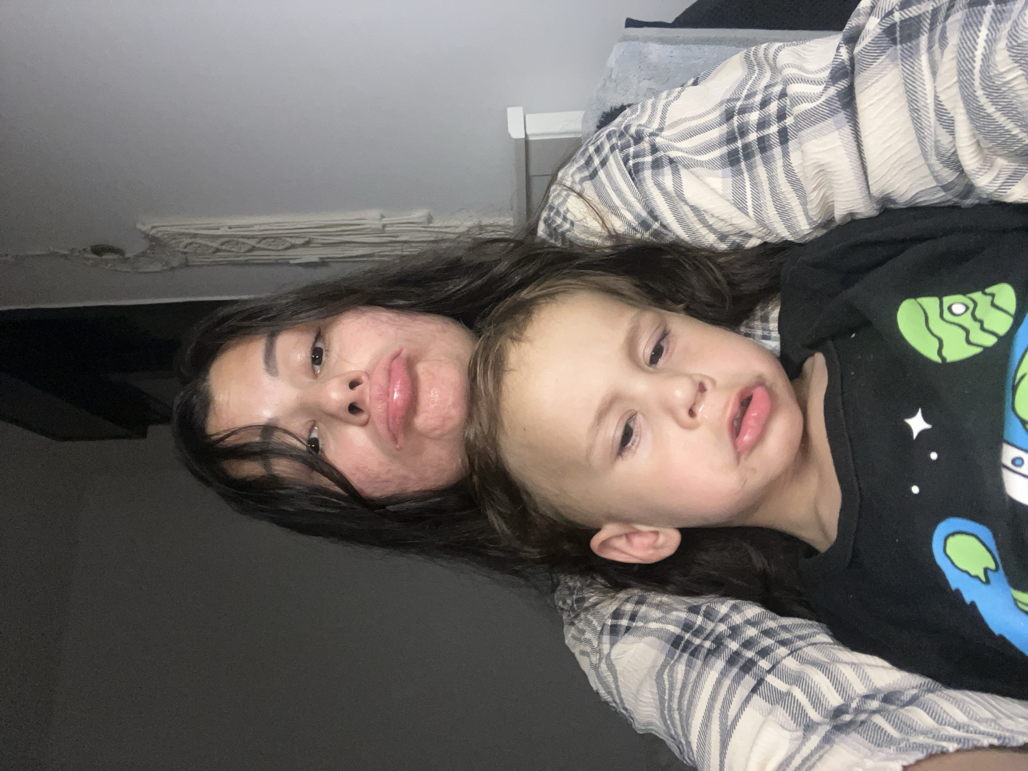 The image presents a medium-close-up, selfie-style indoor shot of an adult female and a young male child. The location of Kiryat Bialik, Israel, cannot be determined from the visual content of the image.
The adult female is positioned behind the child, looking directly at the camera. She has long, dark hair, an oval face, and visible skin texture with some redness on her cheeks and chin. Her lips are heavily pouted and appear glossy. She is wearing a light-colored, possibly cream or off-white, long-sleeved plaid shirt with dark blue or grey lines and a crinkled texture.
The young male child is seated in front of and leaning against the adult female. He has short, light brown hair and a round face, also with pouted lips. His eyes appear heavy-lidded or partially closed and are directed slightly downwards or off-camera. He is wearing a black long-sleeved t-shirt adorned with colorful cartoon-like graphics, including blue, green, and white shapes, and white stars.
Both individuals are making similar pouting expressions. The background on the left side of the frame is a plain dark grey wall. On the right side, a lighter grey wall is visible, featuring a partial view of a light-colored macrame wall hanging and a white or light-toned headboard or furniture element. The overall lighting is dim, suggesting an indoor setting under artificial or low natural light.