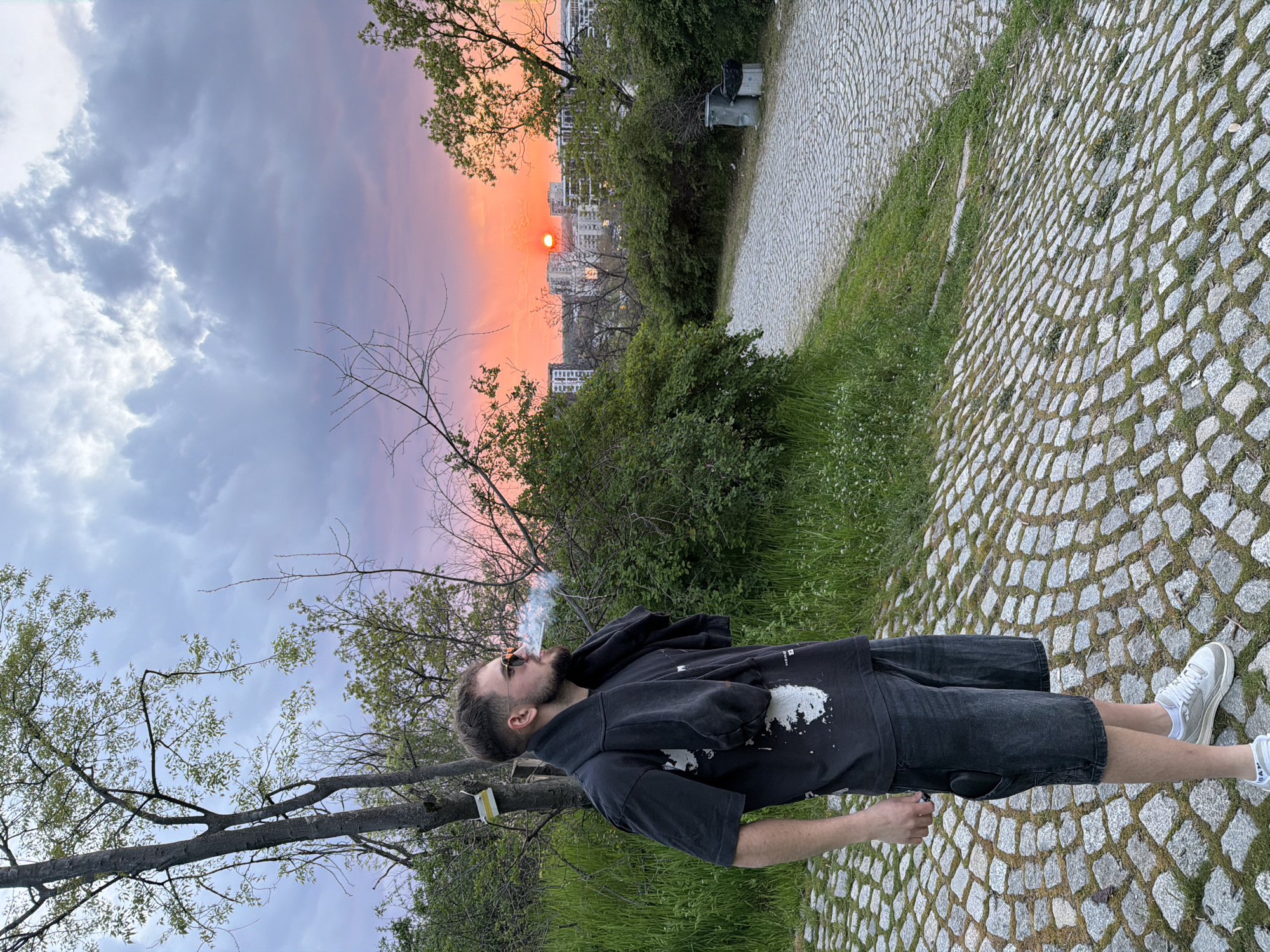 The image shows a man standing on a cobblestone path in a park in Plovdiv, Bulgaria. He is facing away from the camera, looking towards the setting sun. He is wearing a black t-shirt, black shorts, and white sneakers, and is smoking a cigarette or vaping. The sky is a mix of grey clouds and orange and pink hues from the setting sun, with the sun positioned behind buildings on the horizon. Trees and bushes are visible along the path, and in the background, there are some buildings, possibly residential. A public park setting is suggested by the paved path and surrounding greenery. The man is standing near the edge of the cobblestone path, with grassy areas on either side. Trash cans are located in the background.
