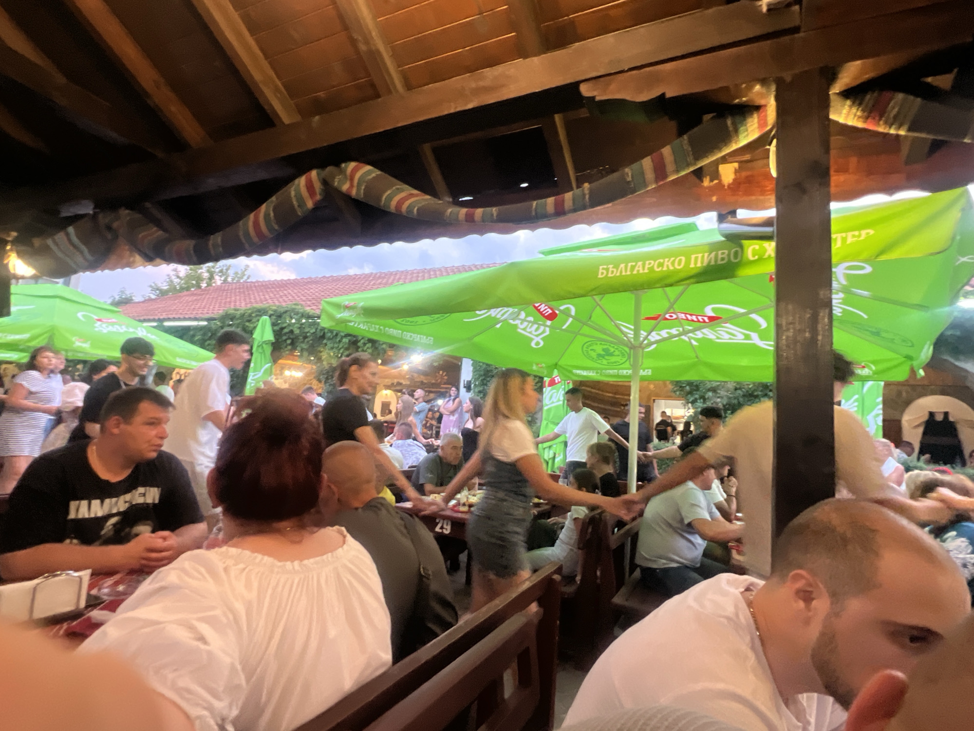 The image depicts an outdoor gathering at what appears to be a restaurant or beer garden. Several people are seated and standing around tables, some of whom are interacting. The scene is covered by a wooden-beamed roof with decorative fabric draped from it. Bright green umbrellas with Bulgarian text, likely advertising a beer brand, are prominently displayed. The lighting suggests it is late afternoon or early evening. The environment has a rustic, open-air feel.