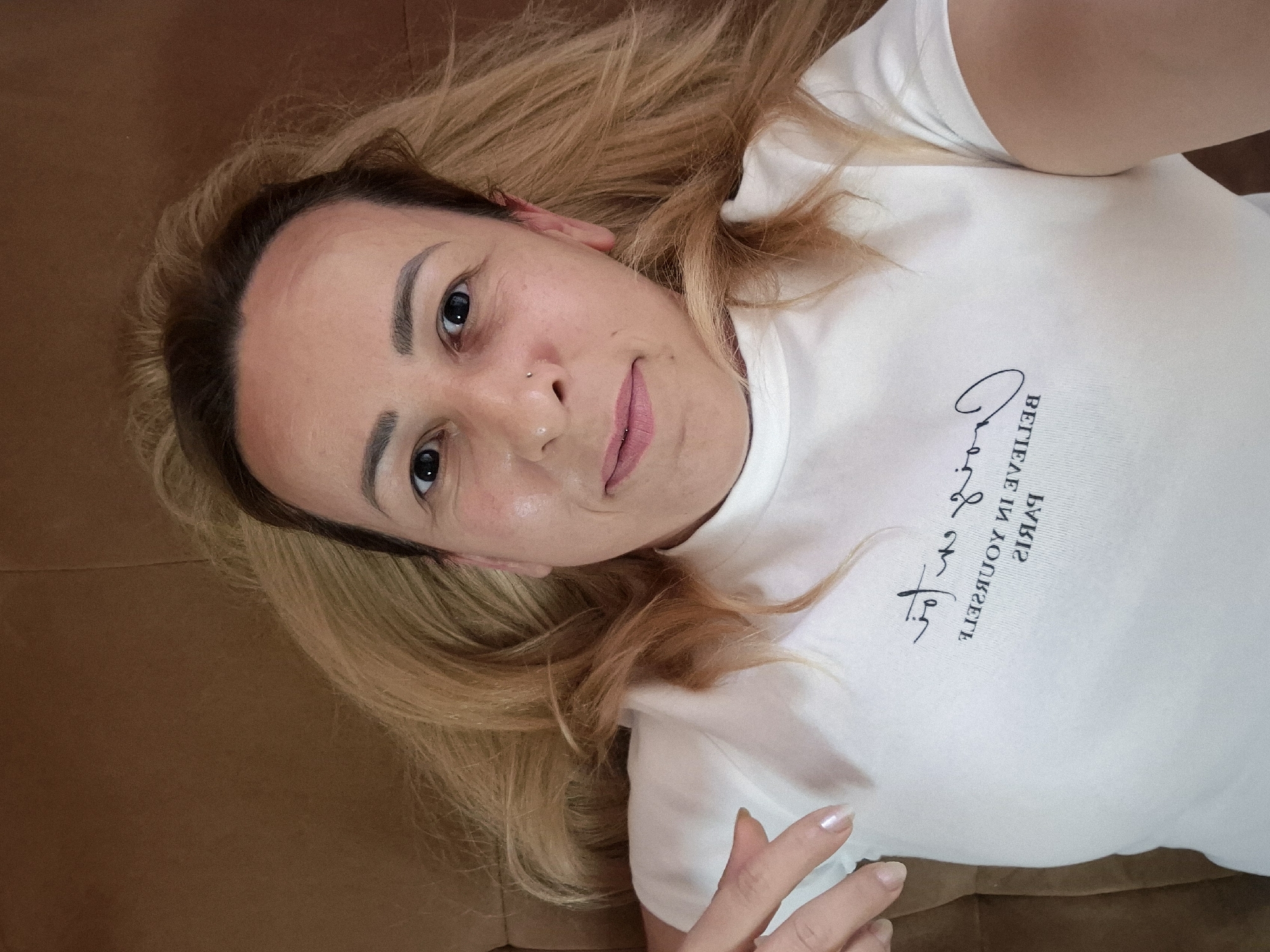 A woman is lying down, possibly on a couch, with her head tilted upwards towards the camera. She has blonde hair, fair skin, and is wearing a white t-shirt with a graphic print. Her left hand is visible, pointing towards her chest. The background is a plain brown surface.