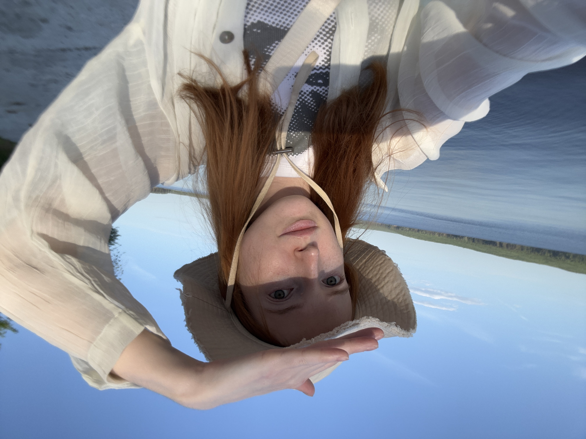 A person with red hair and fair skin, wearing a light-colored hat and a white shirt, is holding their hand up to shield their eyes from the sun. The background shows a body of water and a green shoreline, suggesting a river or lake. The sky is blue. The person appears to be taking a selfie. The image seems to be taken in a natural outdoor setting. The location is the city of Chaikovsky, Russia, based on the context.