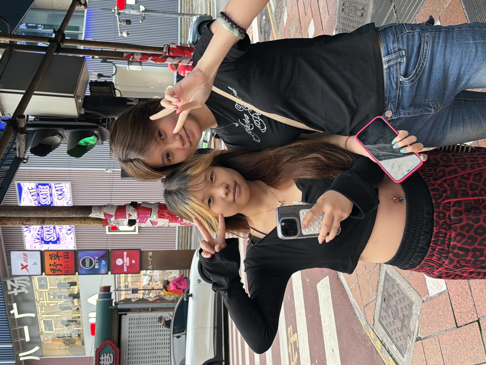 In Yuzhong District, China, two young women are posing for a photo on a city street. The woman on the left, with brown hair with blonde highlights, is wearing a black long-sleeved crop top and red leopard-print pants. She is holding a smartphone in her right hand and making a peace sign with her left. The woman on the right, with short brown hair, is wearing a black t-shirt with a white graphic design and blue jeans. She is also making a peace sign with her right hand and wearing a beaded bracelet on her left wrist. They are standing on a pedestrian crossing with red and white stripes. In the background, there are buildings with various signs, including a traffic light that is currently green. A white car and a darker car are visible on the street. The overall scene depicts a casual, urban setting.