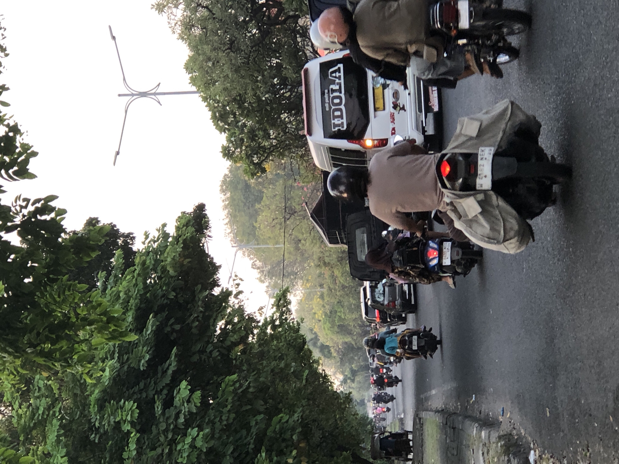 The image captures a busy street scene in Sumorame, Indonesia, likely during a traffic jam. A white van with the word "IDOLA" emblazoned on its rear window is prominently featured, surrounded by numerous motorcycles and a few cars. Several people are riding motorcycles, some with passengers and carrying various items. The road appears to be paved, with a curb on the left side. Lush green trees line the left side of the street, and more trees are visible in the background, suggesting a semi-urban or suburban environment. The sky is overcast, with a street light visible in the upper right quadrant. The overall impression is one of everyday life and transportation in an Indonesian city.
