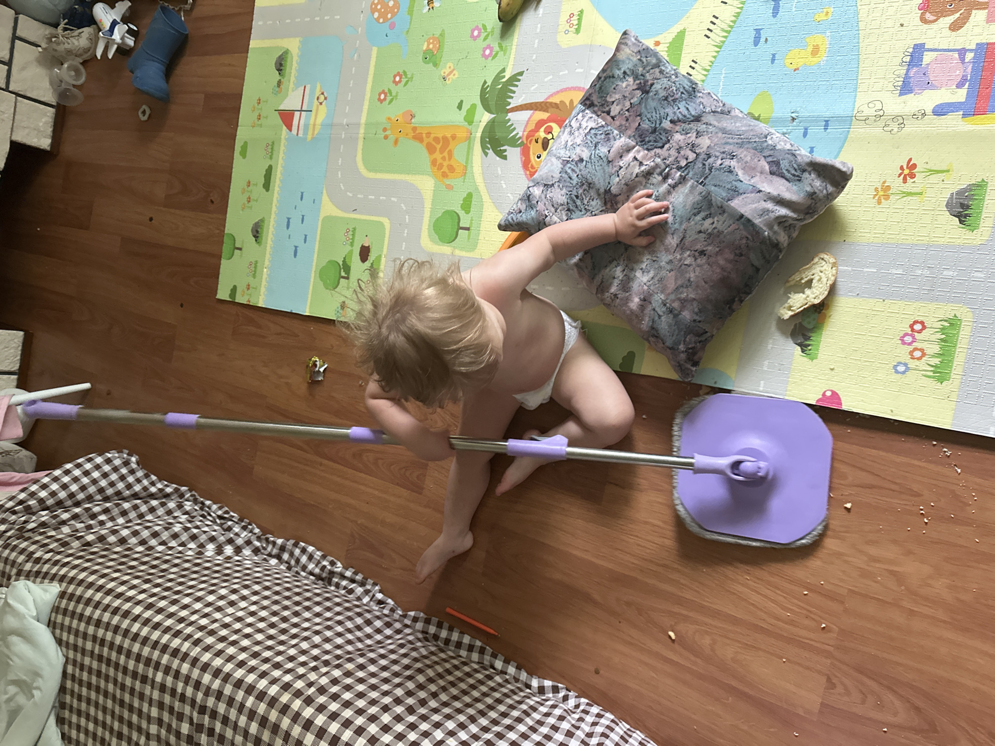 A top-down shot shows a child holding a mop on a wooden floor next to a bed with a brown and white checkered comforter and a colorful playmat. The child, wearing only underwear, sits with their back slightly to the camera and has blonde hair. The playmat, with animal and road illustrations, is located next to a gray patterned pillow. The mop is purple and gray, and its handle stretches toward the bed. A small amount of debris is scattered on the floor. A toy is on the playmat near the top left, and a small orange crayon is on the floor between the child and the bed. There is no information indicating the location in the city of Nizhny Novgorod, Russia.