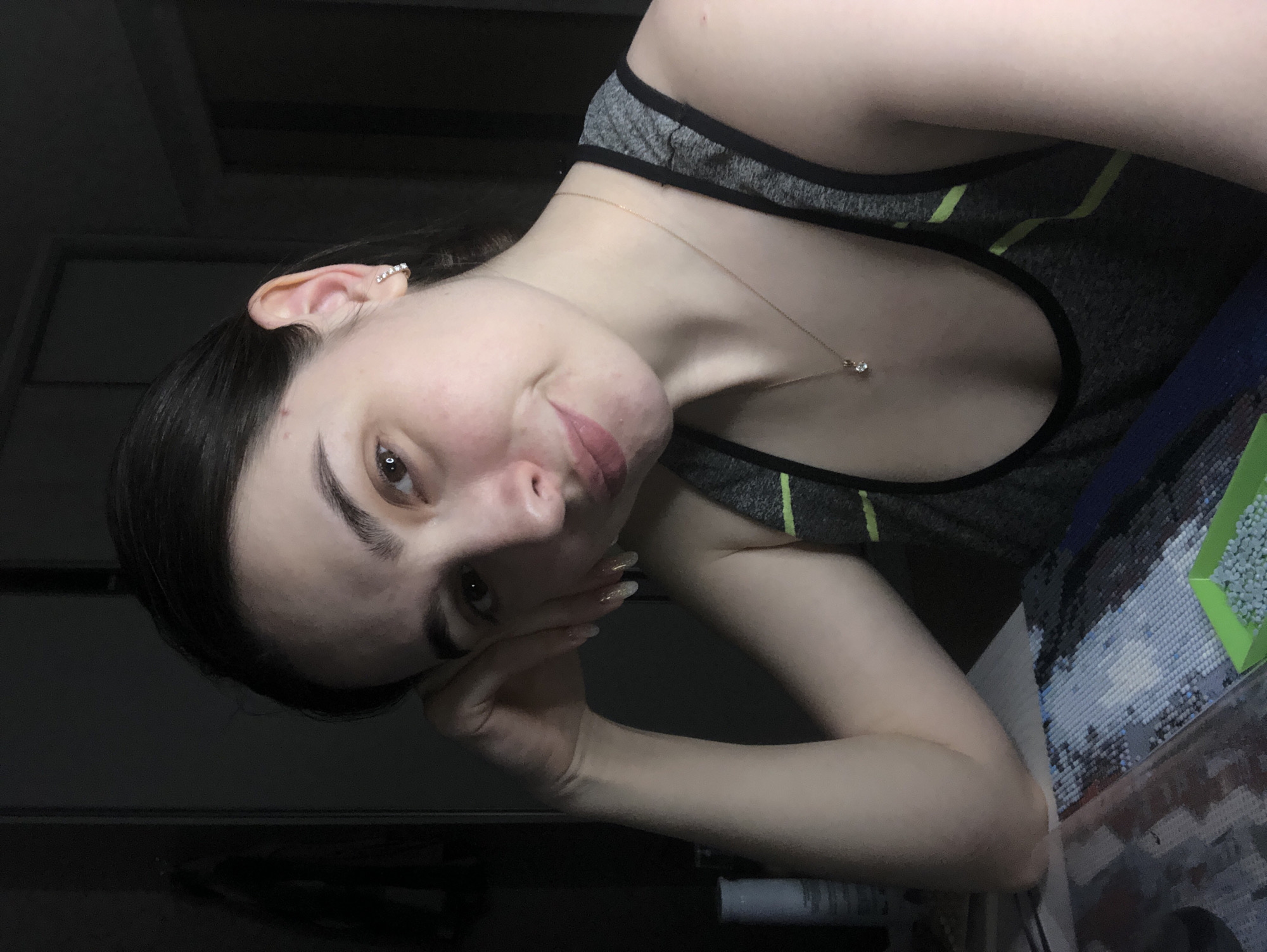 A woman is looking directly at the camera with a slight smile. She is resting her head on her hand. She has dark hair and fair skin and is wearing a gray tank top with black trim and lime green stripes. She has earrings and a necklace. To her right is a diamond painting in progress.
The location in the city of Noviy, Russia, cannot be determined from the image.