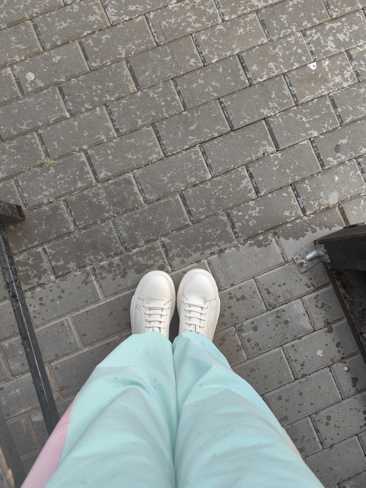 The image shows a person's legs and feet standing on a wet, gray brick pathway. The person is wearing white sneakers and light blue pants with a pink stripe visible on the left leg. The pathway is made of rectangular bricks laid in a stretcher bond pattern. Some of the bricks appear to be damp, with darker patches and lighter, mottled areas. There is a dark metal railing or structure to the left of the frame and a dark wooden structure with a metal bracket to the right. The scene is outdoors, likely in a public space. This setting is consistent with the types of pathways and urban elements found in Myadzyel, Belarus.