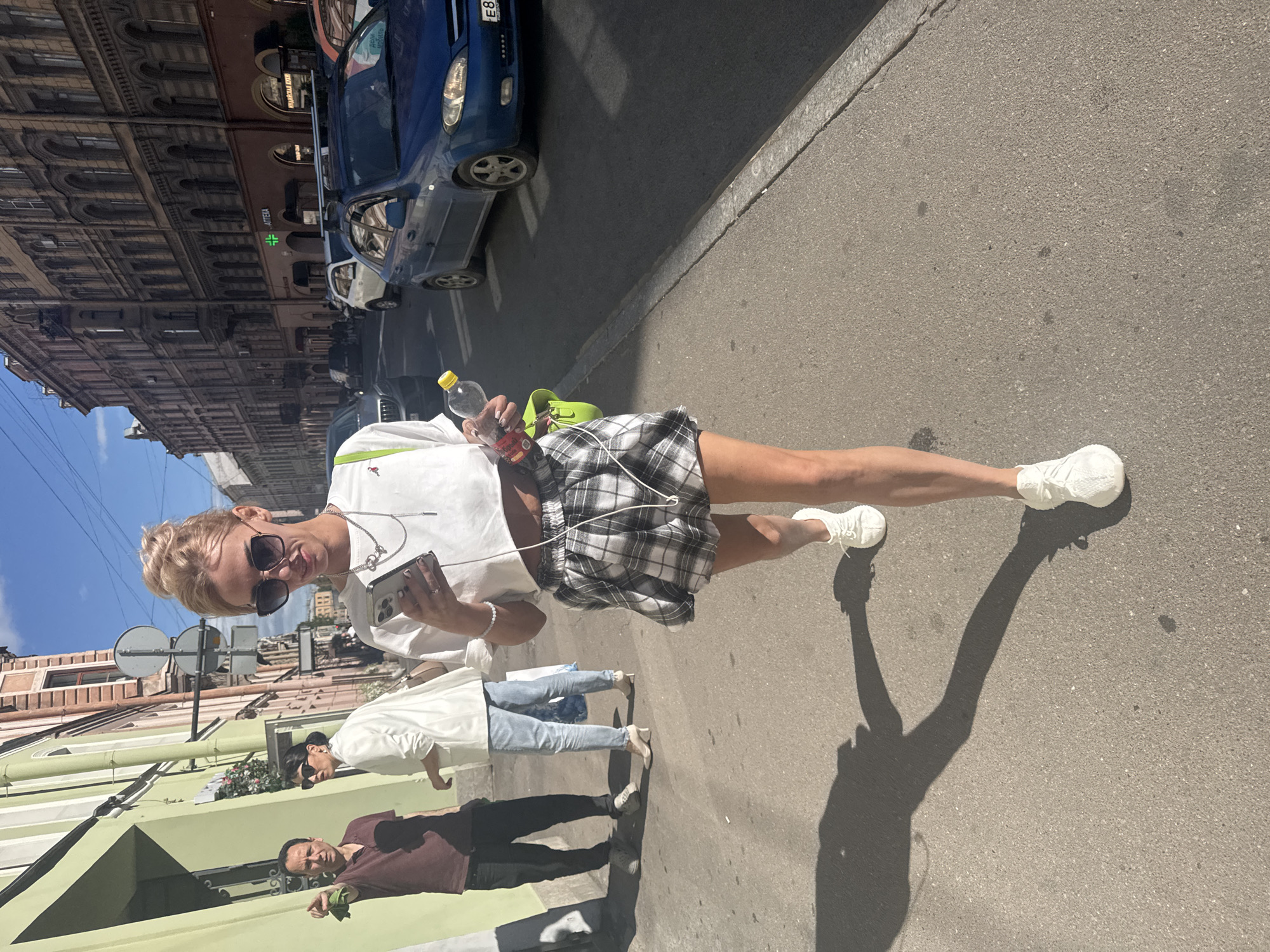 The image depicts a sunny day on a city street in Saint Petersburg, Russia. In the foreground, a young blonde woman with her hair in a bun, wearing large sunglasses, a white cropped t-shirt, and a black and white plaid skirt, is walking and looking at her phone. She is holding a clear plastic bottle with a yellow cap and a bright green purse is slung over her shoulder. Her white sneakers are visible as she walks on the asphalt sidewalk. Behind her, a man in a maroon shirt and dark pants is pointing his left index finger, and two other individuals, one in a white blazer and light blue jeans, and another in a white shirt and light blue jeans, are standing further down the sidewalk. The street is lined with multi-story buildings with arched windows and ornate architecture, characteristic of Saint Petersburg. Several cars are visible on the street, including a blue sedan with a visible license plate. The scene suggests a casual, everyday moment in the urban environment.