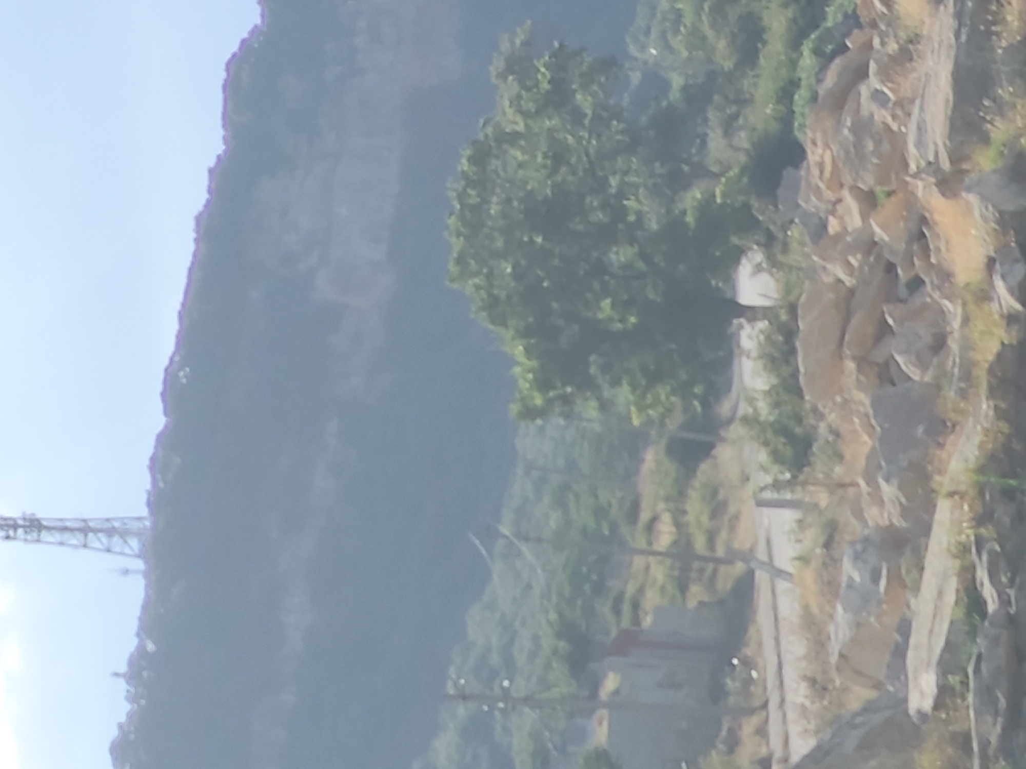 The image shows a hazy landscape with a telecommunications tower on a distant hill. In the foreground, there are rocks and vegetation. A road curves through the scene, and there are power lines and a small structure visible. The overall impression is of a rural or semi-urban environment with a natural backdrop. The image does not provide any specific details that would allow for identification of the location within Makhachkala, Russia, or any particular landmarks.