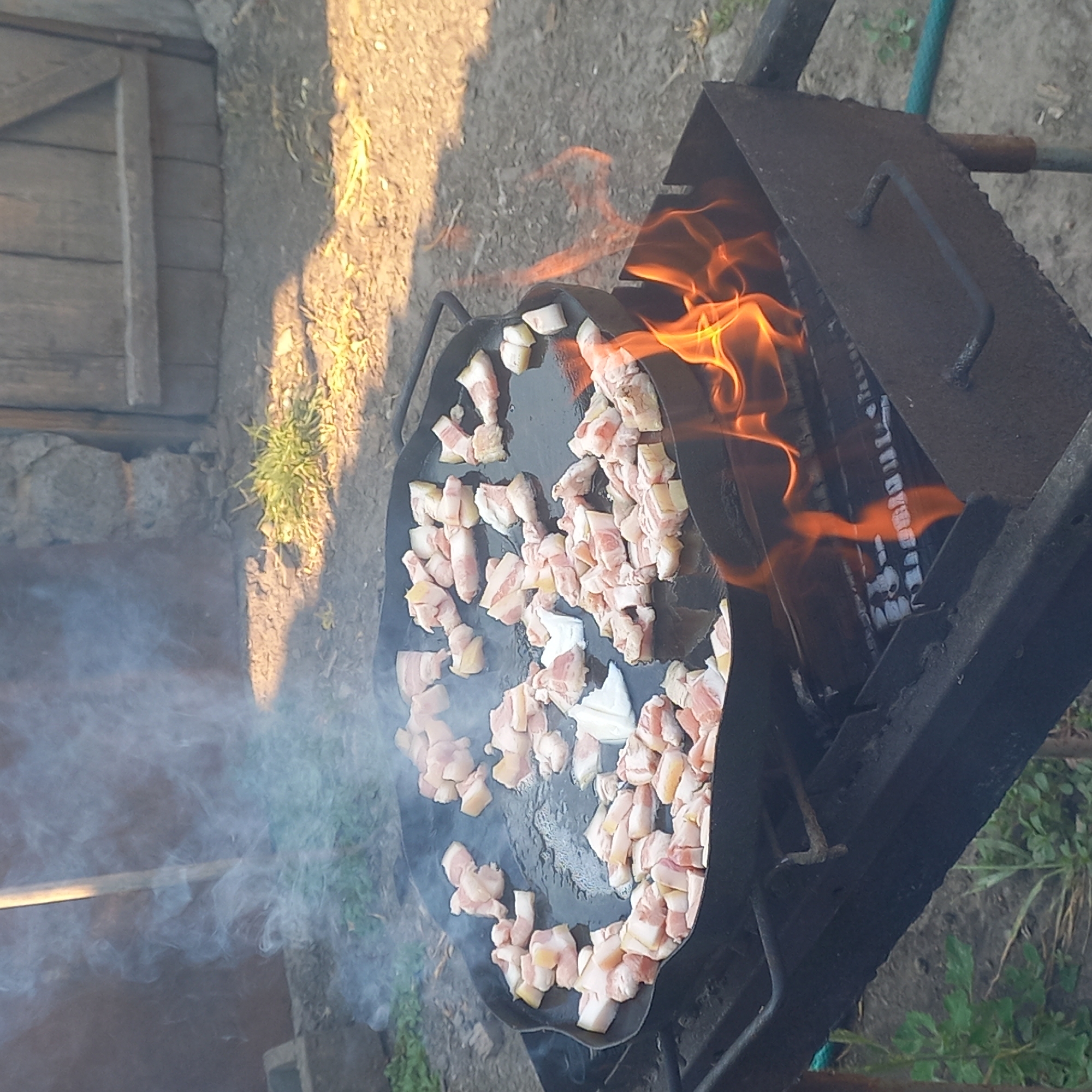 The image depicts an outdoor cooking scene. A large, flat-bottomed, circular metal pan, likely a griddle or plancha, is placed over a charcoal grill. The pan is filled with numerous pieces of chopped raw bacon, which are beginning to render fat and emit smoke. Visible flames from the grill are licking up the sides of the pan, indicating active cooking. The grill itself appears to be a simple, rusted metal brazier with a grate and handles. In the background, the ground is covered with dirt and sparse vegetation, and there is a rustic wooden structure, possibly a fence or part of a building, suggesting a rural or semi-rural setting. The lighting suggests it is daytime, with some shadows cast by the sunlight. There are no people visible in the image. The overall scene suggests a traditional outdoor cooking method, likely for preparing a meal. The question about the location in the city of Klyntsi, Ukraine, cannot be answered from the visual information provided in the image.