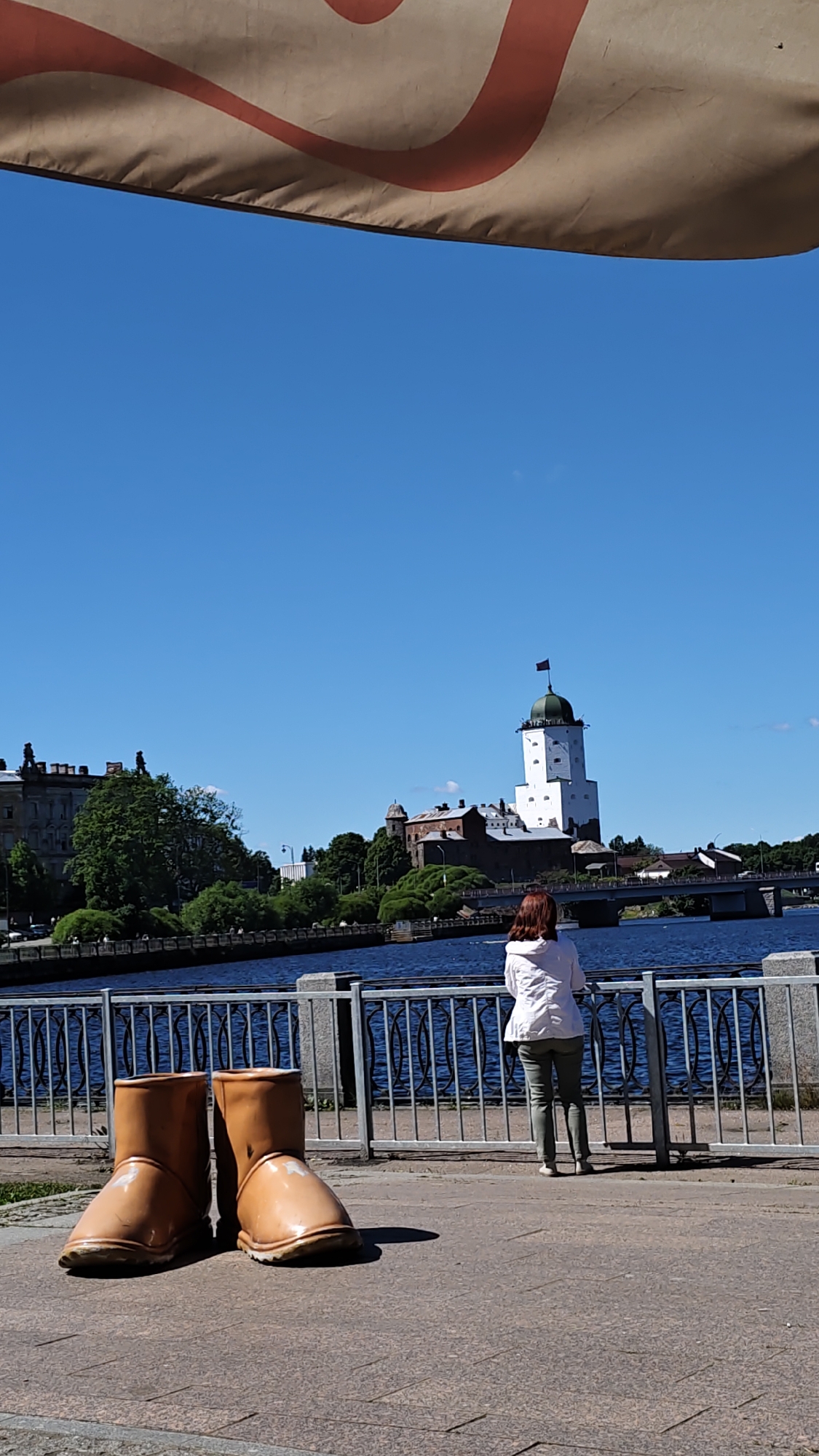 A person with reddish-brown hair, wearing a white jacket and green pants, stands facing away from the camera, looking out at a picturesque waterfront scene. The person is leaning on a metal railing that separates the walkway from a wide, blue river. In the foreground, on the paved walkway, are two oversized, light brown boots, suggesting a quirky art installation or playful display.
Across the river, a white tower with a green-domed roof and a flag at its peak stands prominently. This is likely Vyborg Castle, a historical landmark. Adjacent to the tower are older, darker stone buildings, also appearing to be part of the castle complex. Trees and lush green foliage surround the castle. A bridge spans the river in the middle ground, connecting the two sides.
The sky is a clear, vibrant blue, indicating a sunny day. A partial view of a large, beige canopy with a red curved pattern is visible at the top of the image, suggesting an outdoor seating area or a sunshade. The overall atmosphere is one of leisure and sightseeing on a bright, warm day, possibly during the summer months, in the historic city of Vyborg, Russia. There is no visible text in the image.