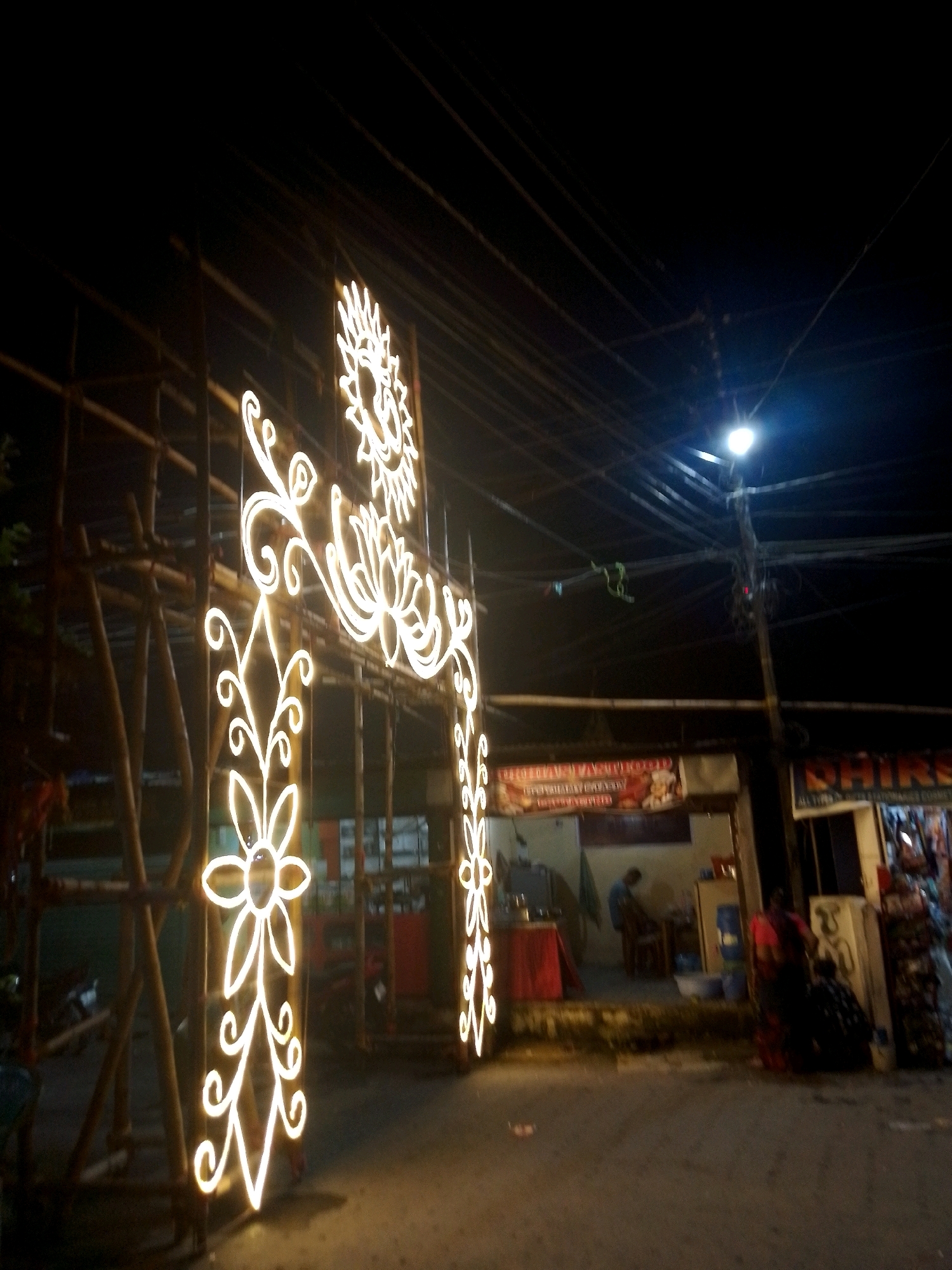 The image, captured at night on a street in Siliguri, India, shows an illuminated festive archway constructed with bamboo scaffolding and adorned with intricate, glowing neon-like lights. The arch features floral motifs and a central sun-like design. In the background, shops are visible, with one displaying a sign that appears to read "CHIRAG." A street light illuminates the overhead electrical wires. Several individuals are present in the scene; one person stands inside a shop, another is seated on the ground near the entrance, and others are seen interacting near the shops. The ground is paved, indicating a public thoroughfare. The overall atmosphere suggests a lively street or market area during a nighttime celebration.