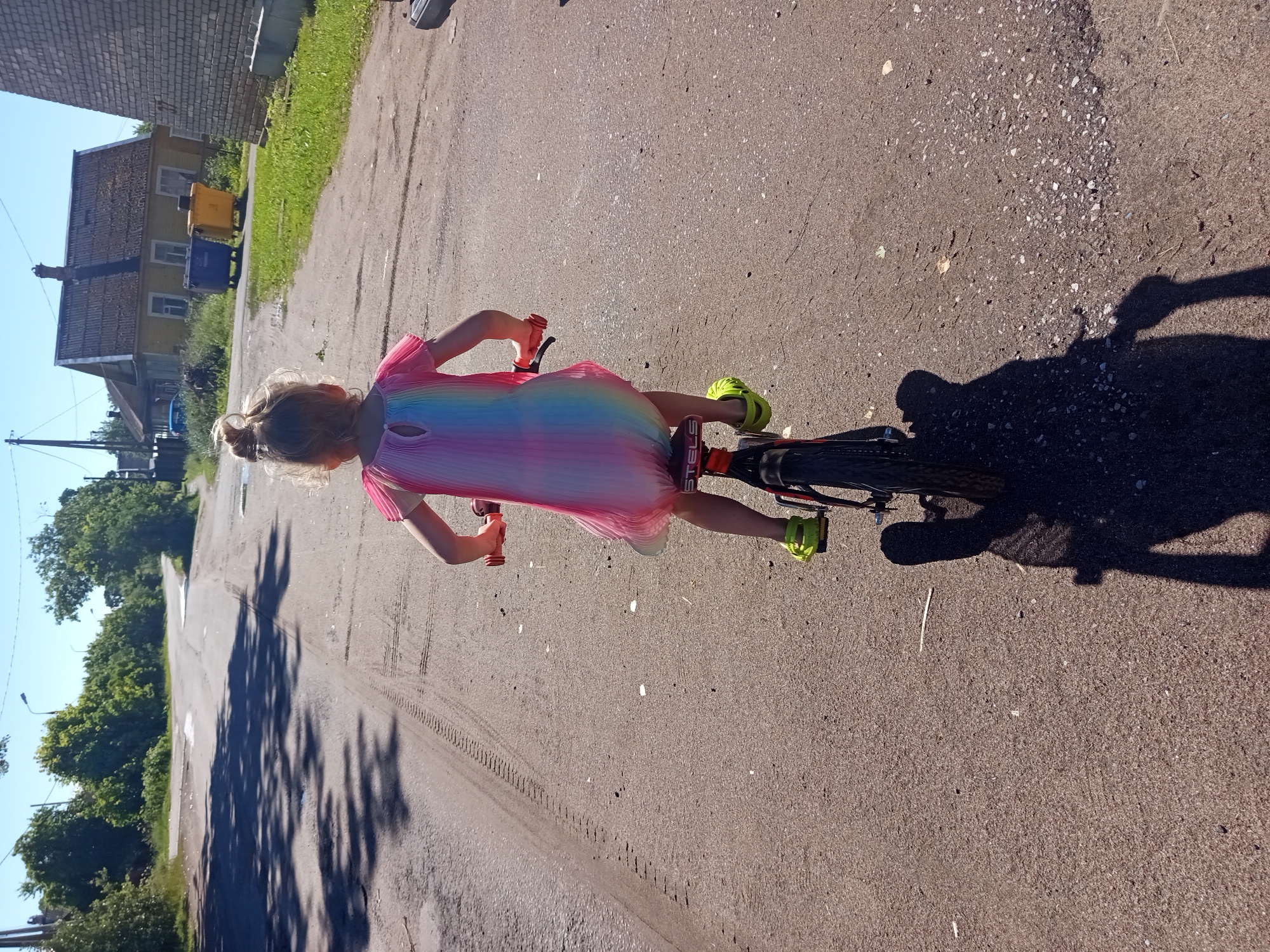 A young girl with blonde hair in a bun, wearing a pink and rainbow-colored dress and bright green shoes, is riding a bicycle on an asphalt road. The girl is facing away from the camera. The bicycle is black with red handlebars and a red seat. The scene appears to be a residential area. In the background, there are houses, trees, and blue and yellow trash bins. The road has tire tracks and shadows of trees are cast across it. The sky is clear and blue. The location is the city of Gdov, Russia.