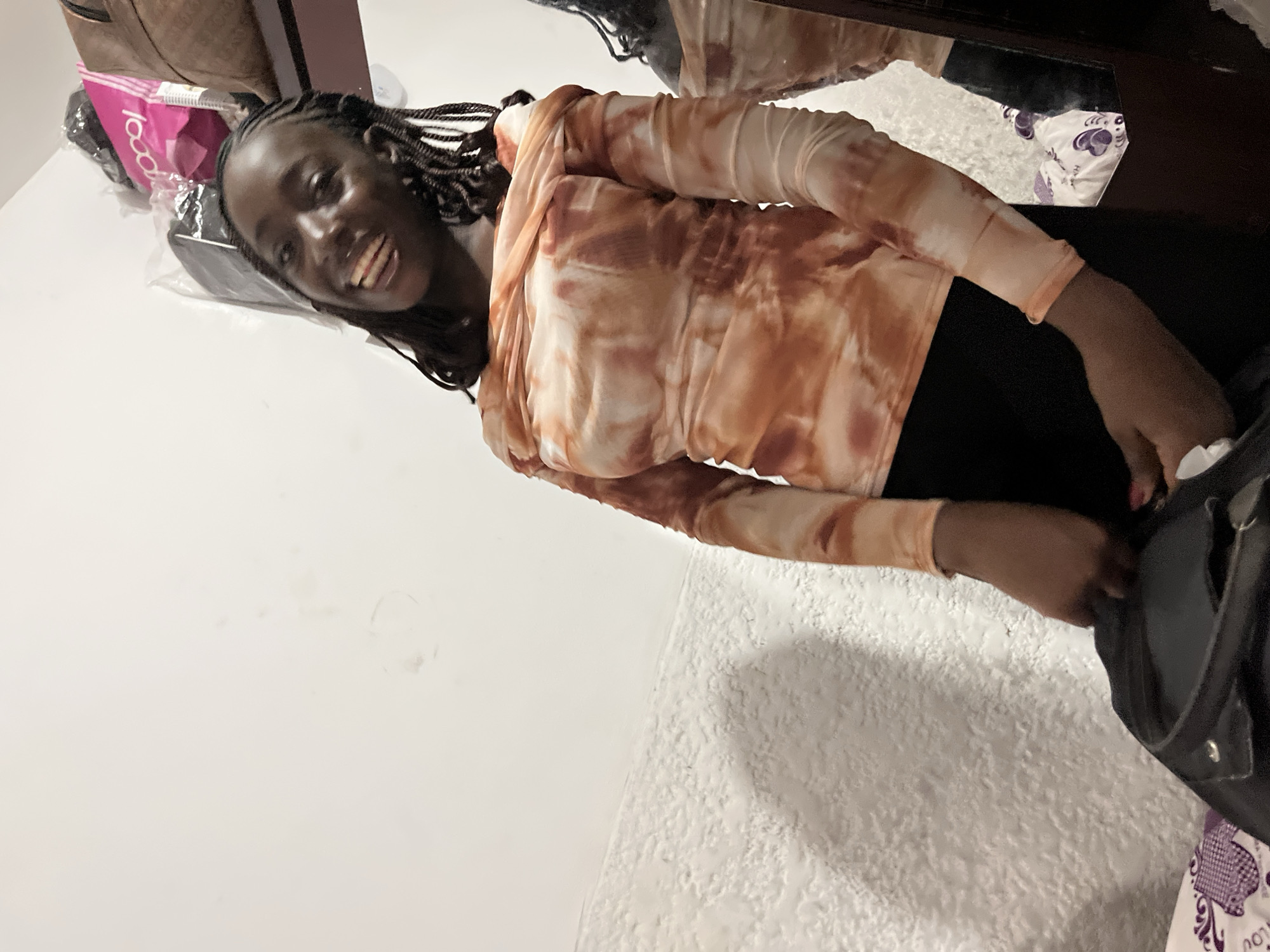 A woman is lying on her side on a white textured surface in a room. She has dark skin and dark hair styled in braids. She is smiling and wearing a long-sleeved shirt with a tie-dye pattern of white and orange. She is also wearing black pants. Her right hand is near a black bag with a silver snap, and there is a white cloth or tissue near her hand. In the background, there is a brown bag, a pink bag with text, and a dark-colored object possibly reflecting in a mirror. The location is possibly a room in Abuja, Nigeria, though there are no specific indicators to confirm this location.