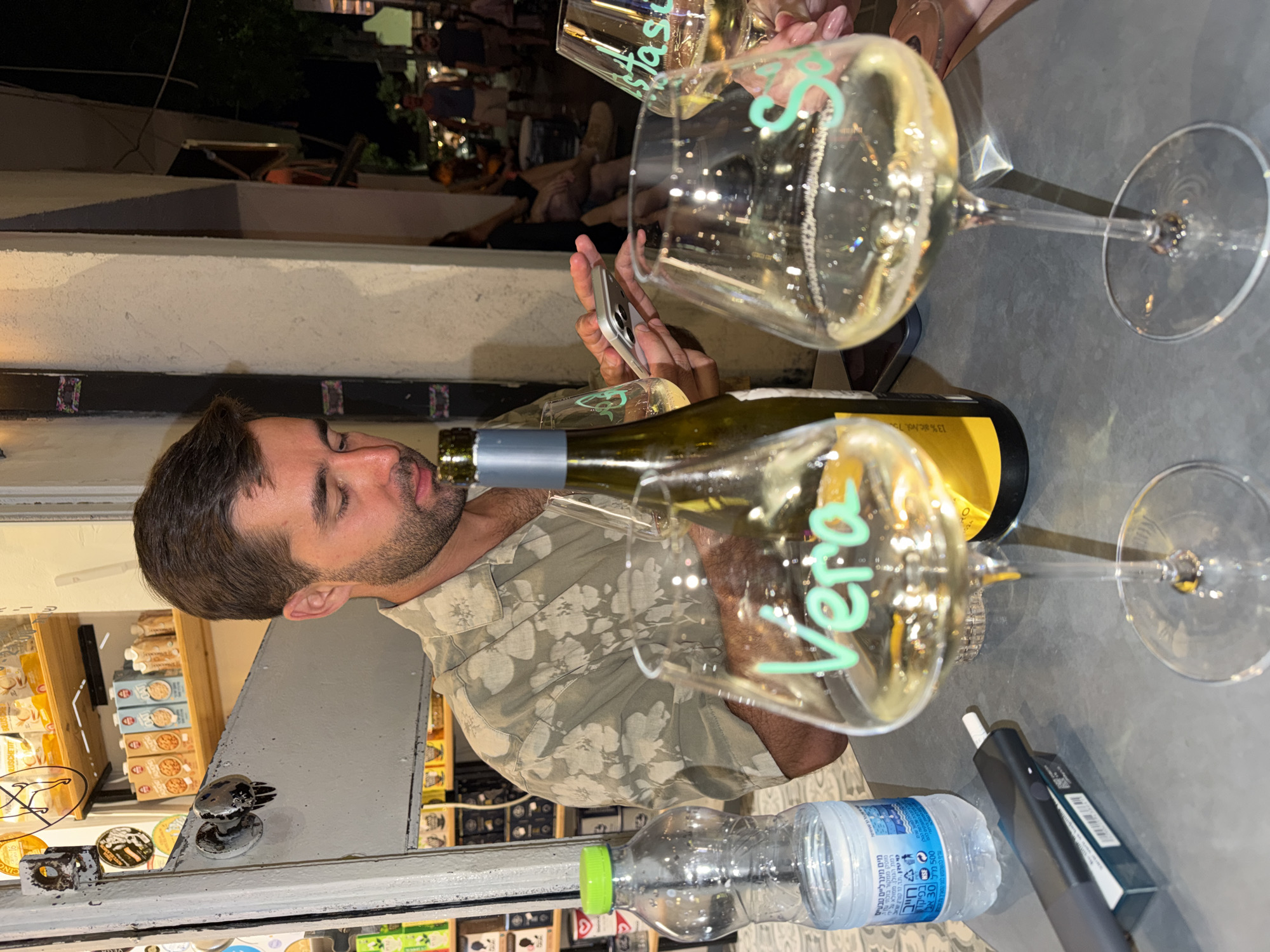 The image captures a candid moment at a street-side establishment in Tel Aviv, Israel, at night. A man, wearing a patterned short-sleeved shirt, is seated at a table and appears engrossed in his mobile phone, which he holds in his hands. In front of him, two wine glasses are prominent. The glass on the left has the name "Vera" written in green on its surface. The glass on the right has "Sasa" written in green. A bottle of white wine, with a silver cap and a yellow label, stands between the glasses. A plastic water bottle with a green cap is also visible on the table, along with a sleek, dark electronic smoking device and its packaging. The background reveals shelves stocked with various food products, suggesting the establishment might be a shop or a cafe. The overall ambiance is relaxed and casual, indicative of an evening out in the city.