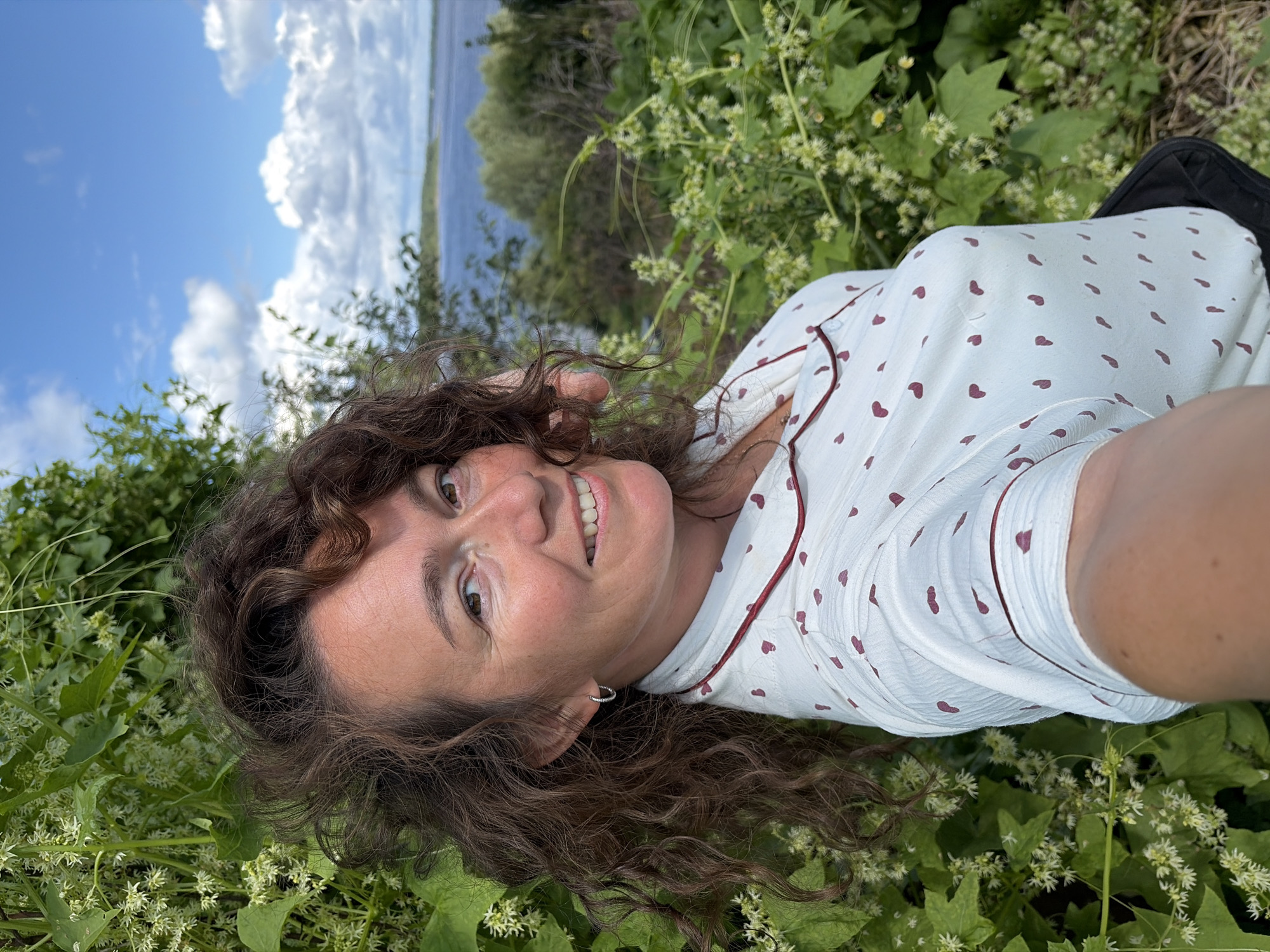 A woman with curly brown hair and fair skin is taking a selfie outdoors, with a body of water and a cloudy blue sky in the background. She is wearing a white shirt with a pattern of small maroon hearts and a maroon trim around the collar and sleeves. A silver hoop earring is visible in her left ear. She is smiling and her head is tilted slightly to her right. Lush green foliage with small white flowers surrounds her on the left and in the foreground. The location is the village of Olkhovka, Russia.