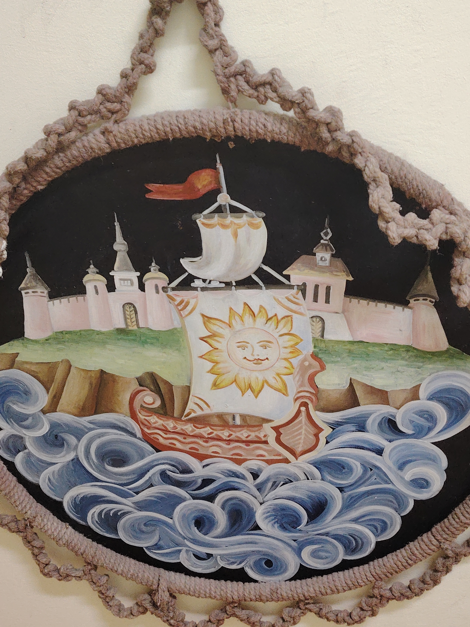 The image is a decorative artwork depicting a ship with a sun on its sail, sailing on a turbulent sea. Behind the ship is a fortress or castle built on a cliff. The artwork is framed with a beige macrame rope. The scene is painted in a folk art style, with bold outlines and stylized elements. There is no indication of Sarapul, Russia, in the image.