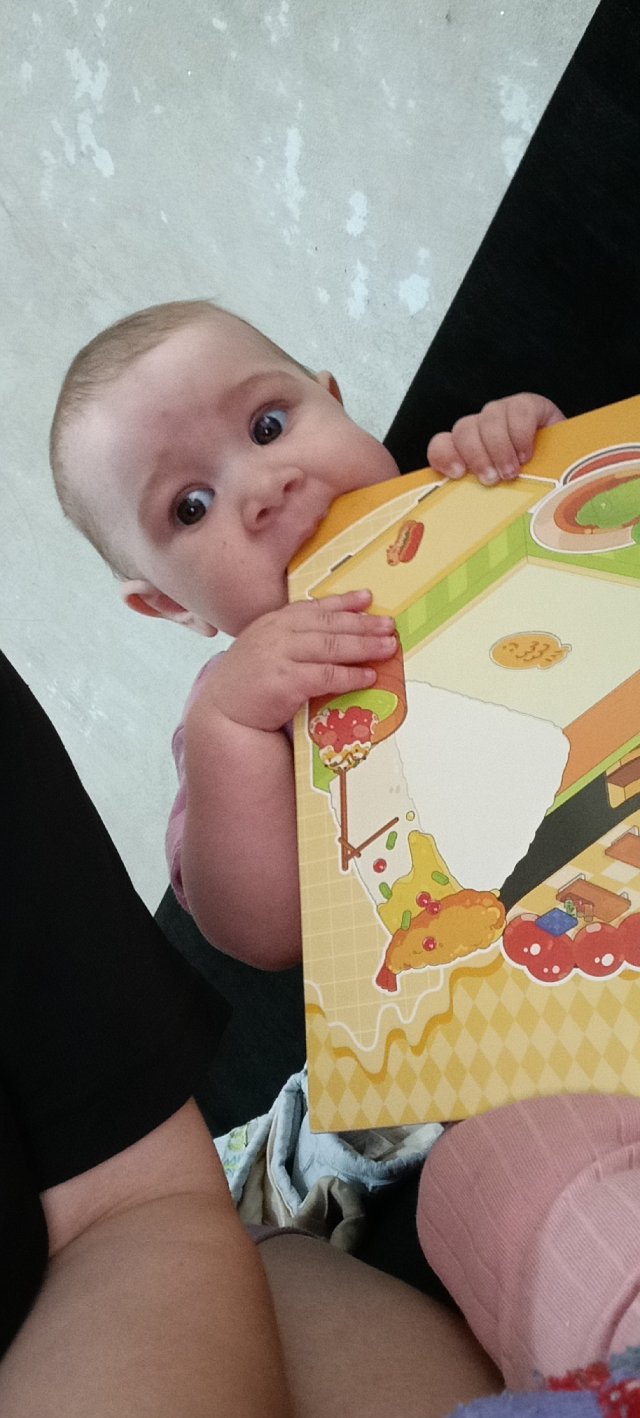 A baby is biting a book. The baby has light skin and light brown hair. The baby is wearing a pink shirt. The book has a colorful illustration of food. The baby is being held by an adult, whose legs are visible in the foreground. The background is a light-colored wall. The scene appears to be indoors. The location in the city of Хохлы, Russia is not discernible from the image.