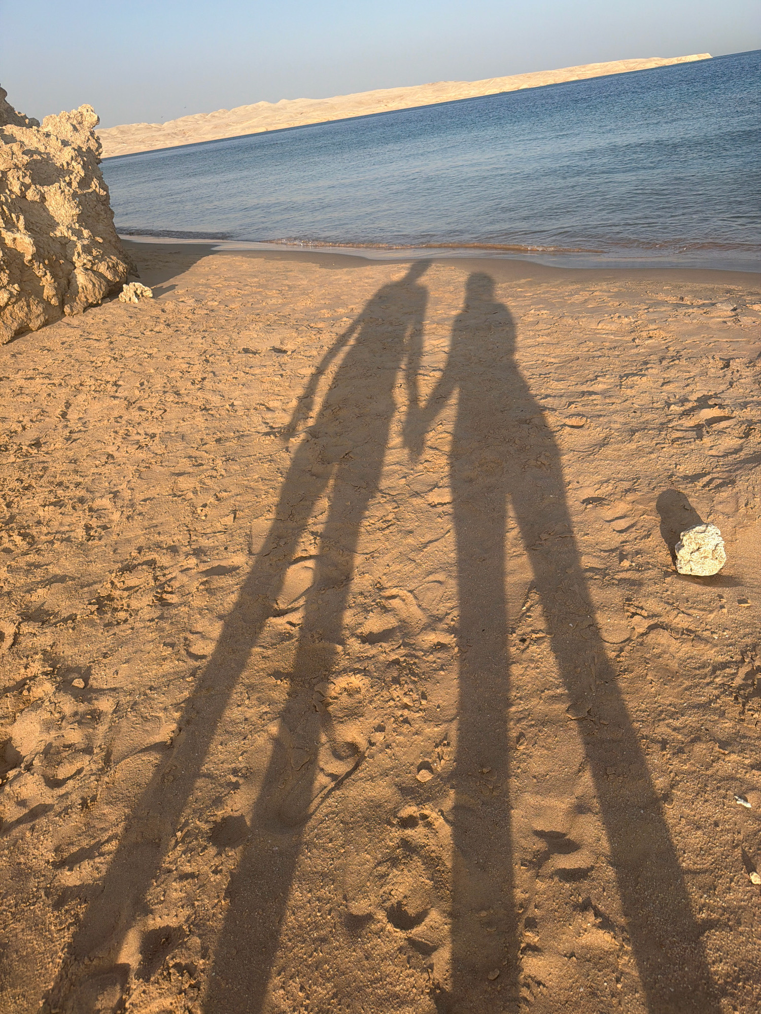 The image depicts a serene beach scene at sunset in Sharm El Sheikh, Egypt. The foreground is dominated by the sandy beach, casting long, dramatic shadows of two people holding hands. The sand is textured with ripples and a few scattered stones, one of which is clearly visible to the right of the shadows. In the background, the calm, blue waters of the sea gently meet the shore. Beyond the water, a distant coastline with undulating, sandy hills stretches across the horizon under a clear, pale blue sky. The overall atmosphere is peaceful and romantic, suggesting a shared moment between two individuals.