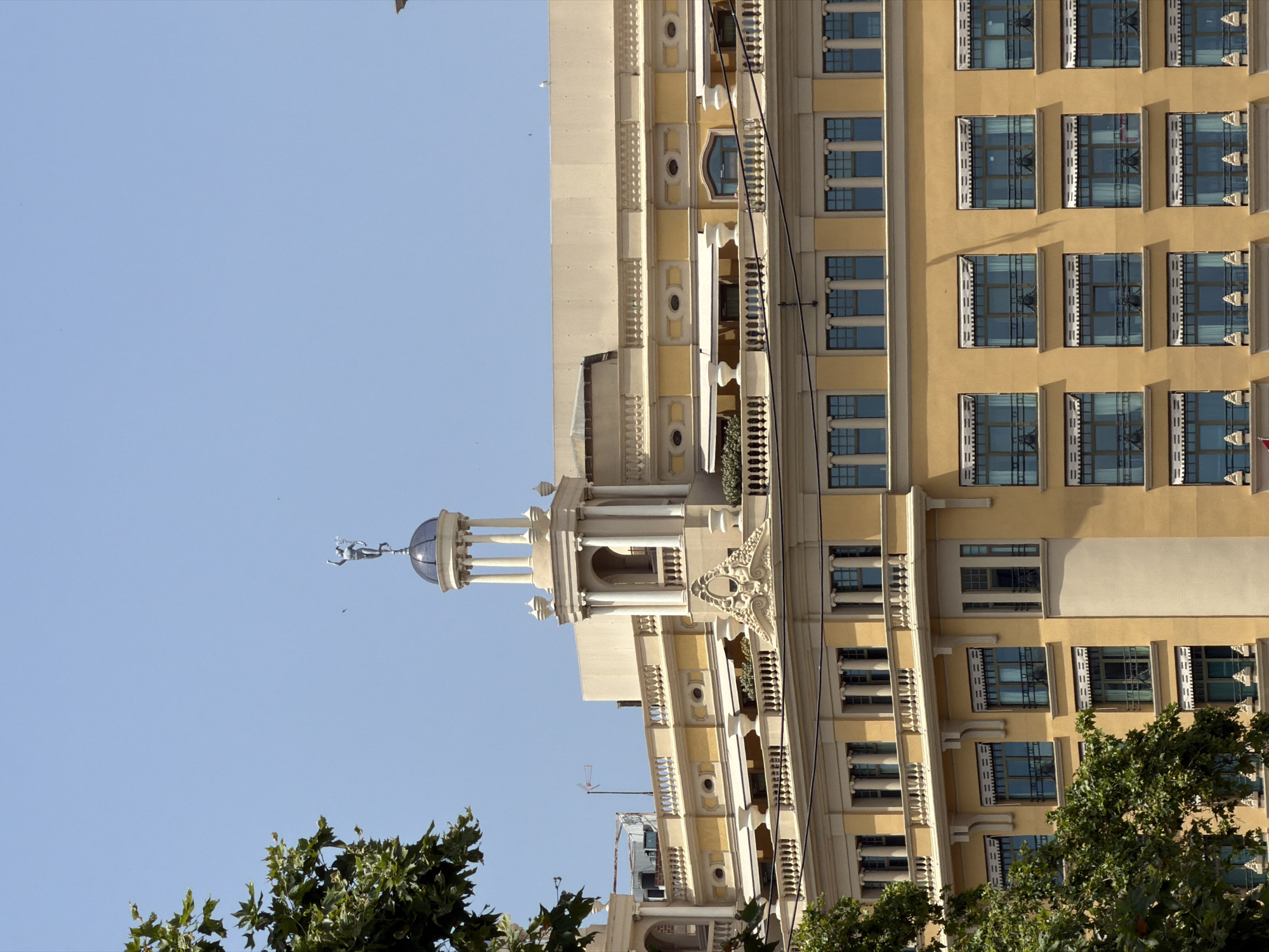 The image shows the facade of a large, light yellow building in Barcelona, Spain. The building has a classical architectural style with ornate details, including columns, balustrades, and a central dome topped with a statue. Several rows of windows, many with awnings, are visible on the facade. A large tree with green leaves partially obscures the left side of the building. The sky is clear and blue. The image appears to be taken during the daytime.