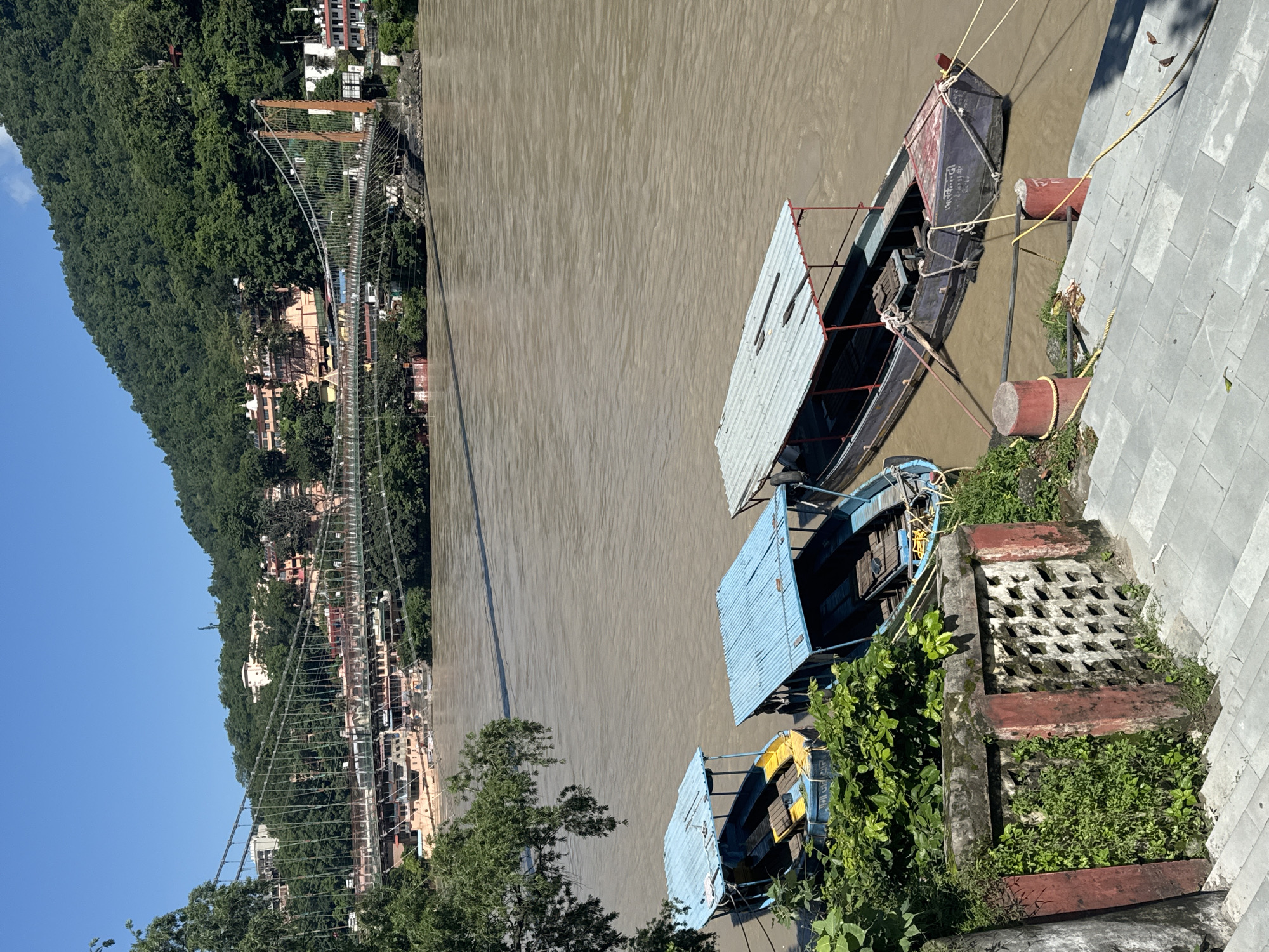 This image shows the Lakshman Jhula suspension bridge in Rishikesh, India. The bridge spans a wide, muddy river, with lush green hills rising behind it. Several boats are docked along the riverbank in the foreground, some with blue roofs and others with grey. A stone wall with red accents and intricate patterns separates the boats from a set of stairs leading up from the river. The sky is clear and blue, indicating a sunny day. Buildings are visible on the hillsides on either side of the river, suggesting a populated area. There are no people clearly visible in the image.