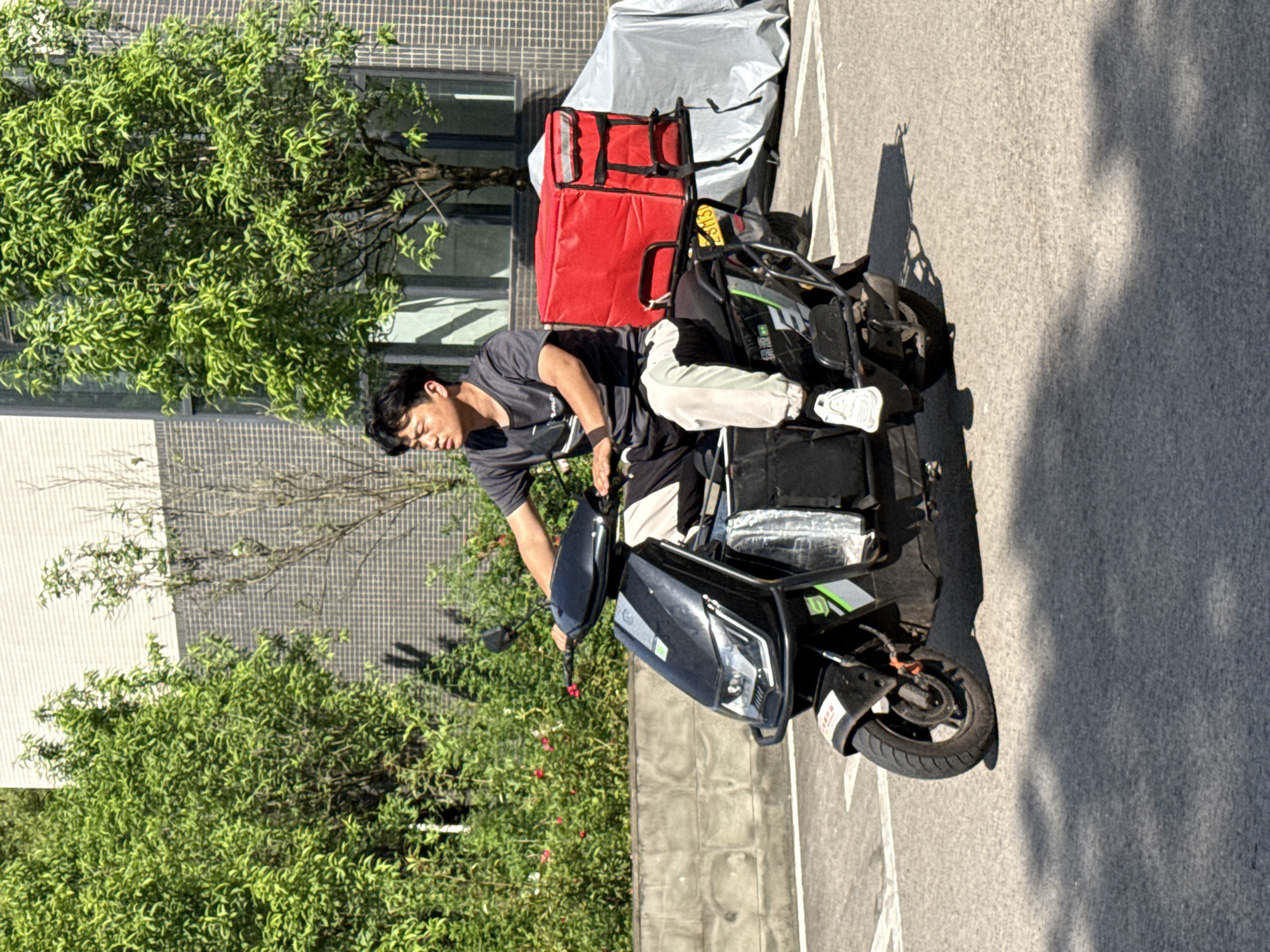 An outdoor scene captures a young man, likely of East Asian descent, resting or sleeping on a black electric scooter. He is dressed in a dark grey t-shirt, light-colored sweatpants, and white sneakers. His head is tilted, and his eyes are closed, indicating a moment of repose, possibly due to fatigue from his work.
The scooter is equipped with a large, bright red insulated delivery bag on its rear rack, suggesting the man is a food or package delivery driver. A smaller black bag hangs from the handlebars. The scooter itself has some green accents, including a stylized "D" or similar letter on its front side panel. White painted lines are visible on the asphalt ground where the scooter is parked.
In the background, a building with a grey tiled facade and windows is visible, along with lush green trees and foliage on both sides of the frame. Strong sunlight casts distinct shadows on the ground, indicating a clear, sunny day, likely morning or late afternoon. The overall setting appears to be an urban or suburban area, possibly a parking lot or a street in Jiangjin, China, consistent with the widespread use of electric scooters for delivery in the region. Visible text includes a partial "2B M3" on the red delivery bag, and some indecipherable characters on a white label on the front fender.