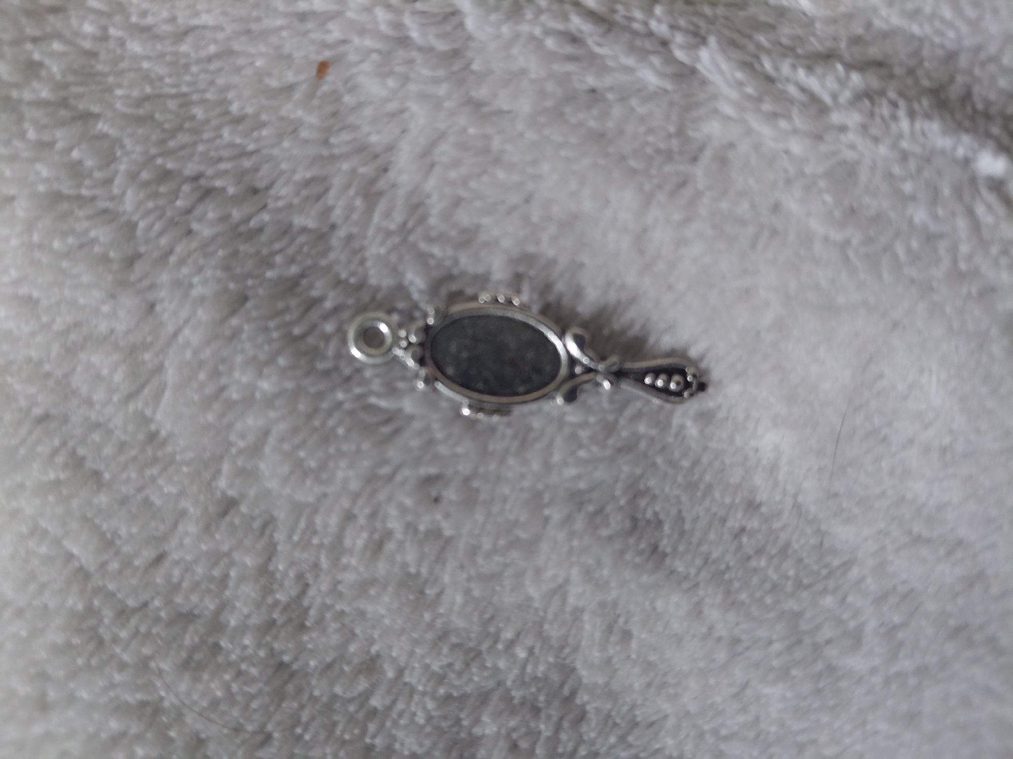 A silver-toned metal charm, designed as an antique handheld mirror, rests slightly right of center on a soft, textured, light-colored fabric surface. The charm features an oval 'mirror' section that appears dark and non-reflective, encased within a decorative, raised metal frame. A slender, ornate handle, adorned with small raised dots, extends downwards from the base of the mirror frame. At the top of the charm, a small, circular loop provides an attachment point. The underlying fabric is off-white or light gray with a visible, shaggy pile, indicative of a blanket or plush textile. A thin, dark, curved hair is present on the fabric in the upper left portion of the image, and a small, reddish-brown speck is visible on the fabric to the right of the charm. The scene is illuminated by soft, diffuse light. No individuals, specific actions, or identifiable geographical markers of the city of Kyiv, Ukraine, are present in the image.