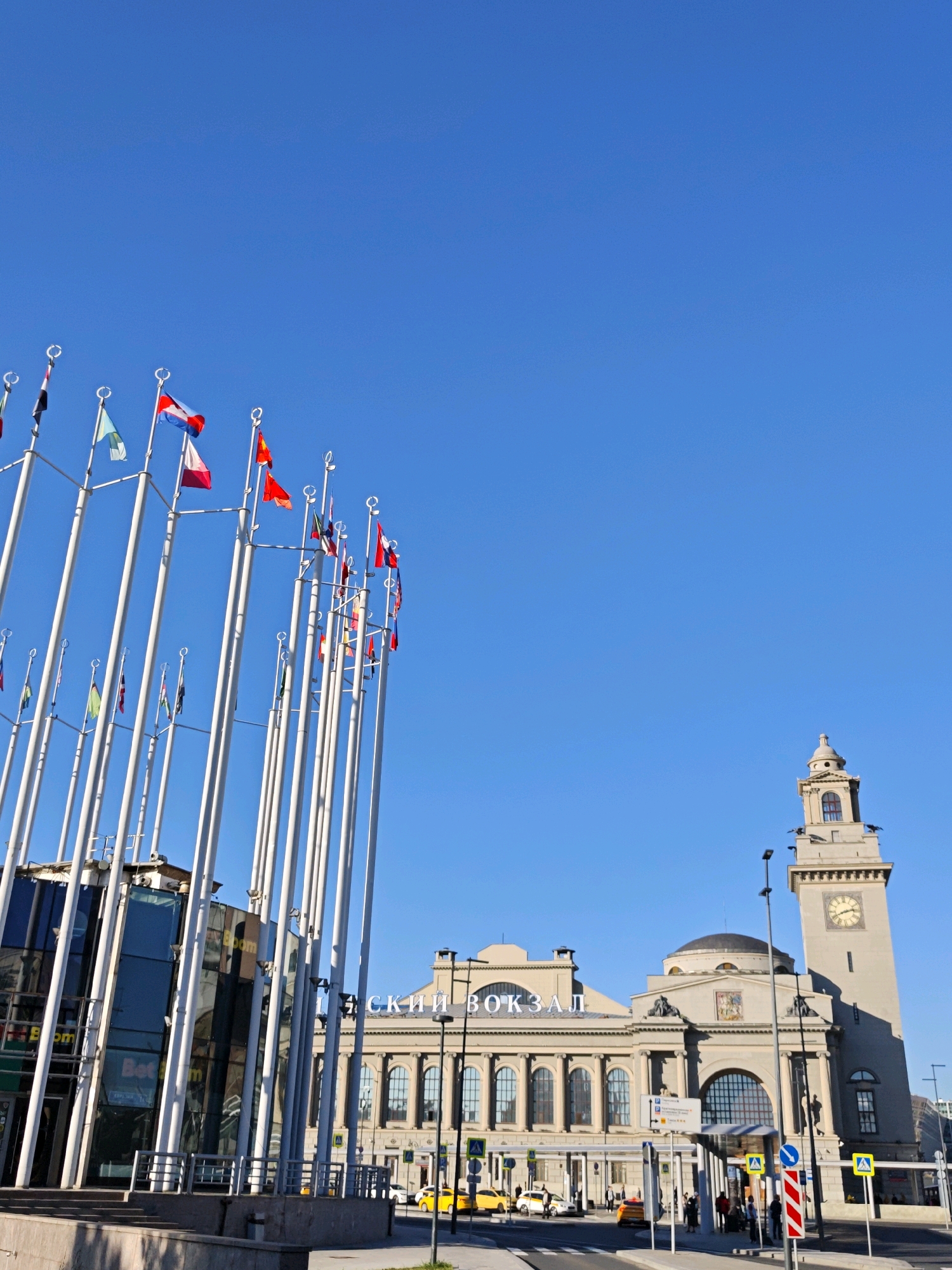 The image depicts the Leningradsky Railway Station in Moscow, Russia, on a clear, sunny day. In the foreground, a cluster of tall, white flagpoles stands to the left, adorned with various national flags, suggesting an international or diplomatic setting. To the right of the flagpoles, a modern glass building with "BetBoom" signage is visible.
Dominating the background is the grand facade of Leningradsky Railway Station, characterized by its neoclassical architecture, featuring arched windows, columns, and a prominent dome. A tall clock tower with a visible clock face rises from the right side of the station building.
The street in front of the station is active with several yellow taxis and a few pedestrians walking. Road signs and pedestrian crossings are visible, indicating a typical urban street environment. The sky is a vibrant, cloudless blue, casting clear shadows and highlighting the architectural details of the buildings and the flags.